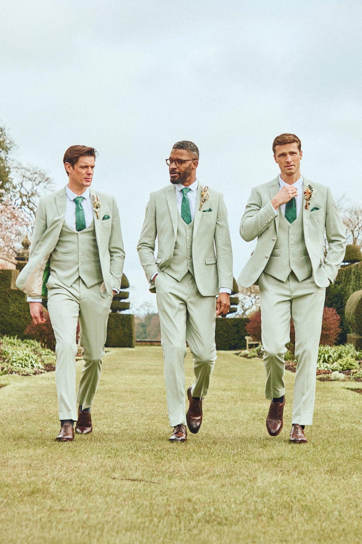 Three men in Marc Darcy’s FINLEY - Moss Green Three Piece Suit, complete with matching ties and boutonnieres, walk outdoors on a manicured lawn—ideal summer wedding attire.