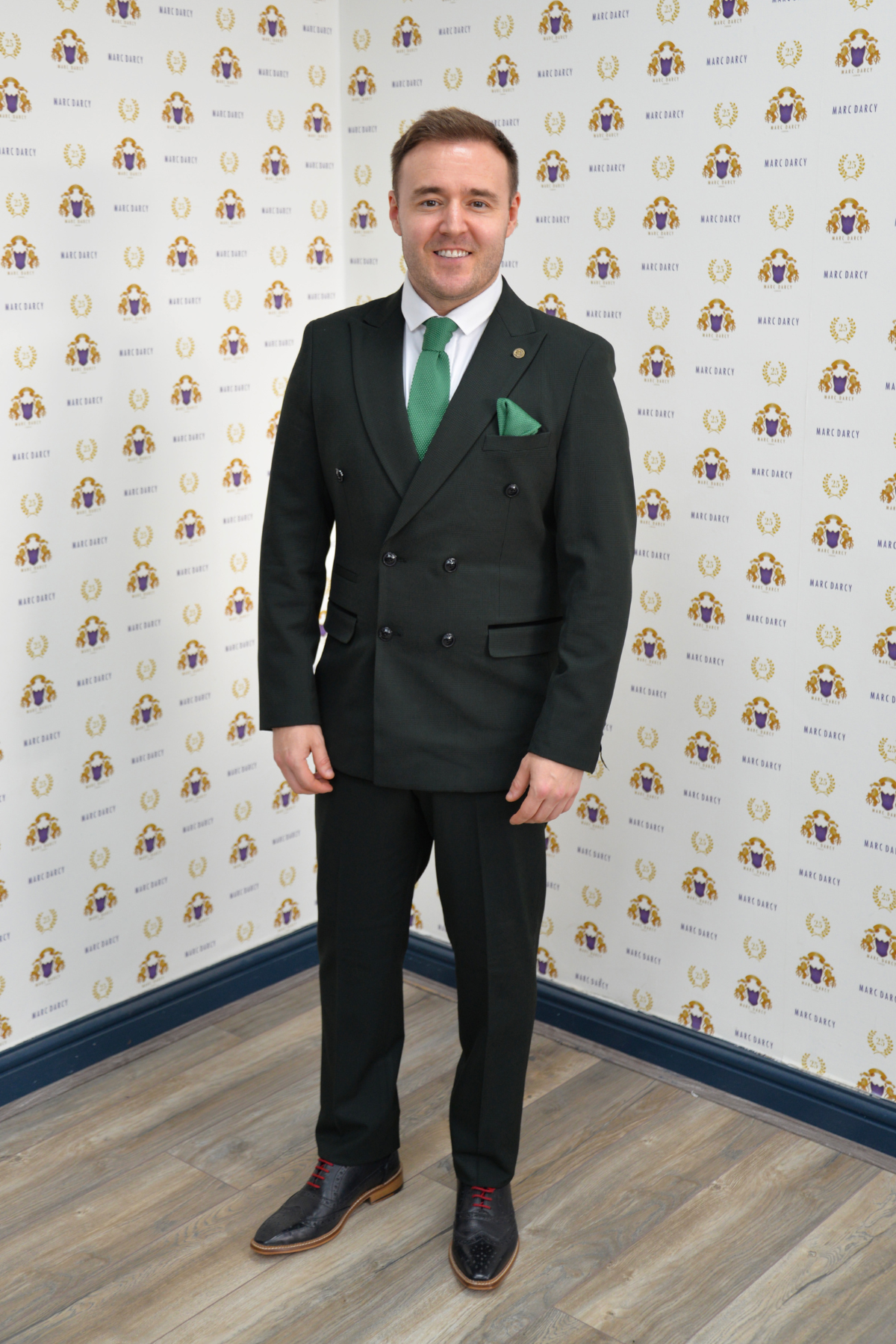 Coronation Street’s Alan Halsall (Tyrone Dobbs) wears a Marc Darcy BROMLEY olive green double-breasted suit, white shirt, and green tie as he smiles in front of a step-and-repeat banner with yellow, purple, and gold logos on white.