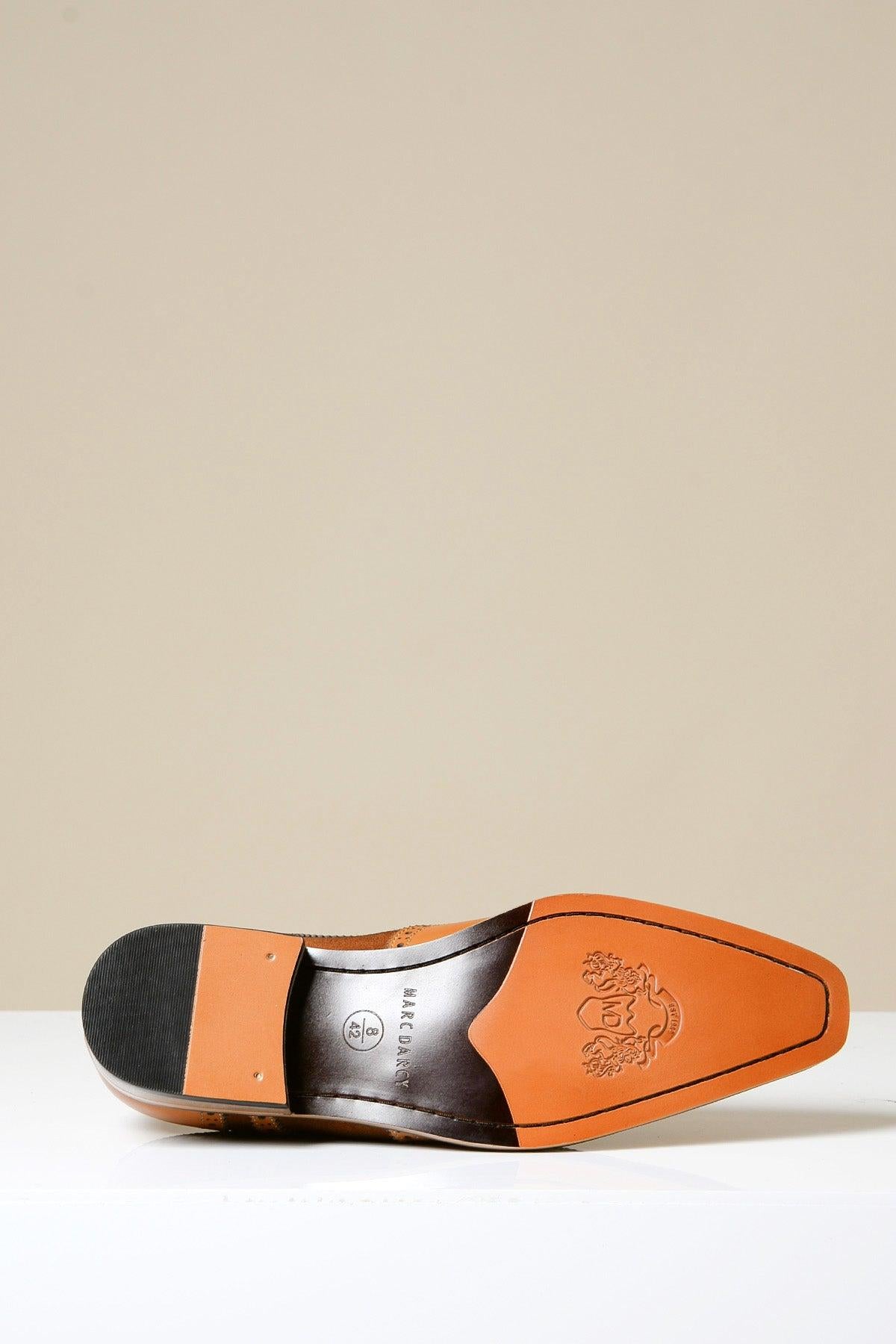 The BRADLEY Tan Leather Suede Contrast Brogue Shoe by Marc Darcy is displayed sole-up, featuring a pointed toe, light brown full grain leather soles, black heel cap, and embossed lion crest near the toe on a plain beige background.