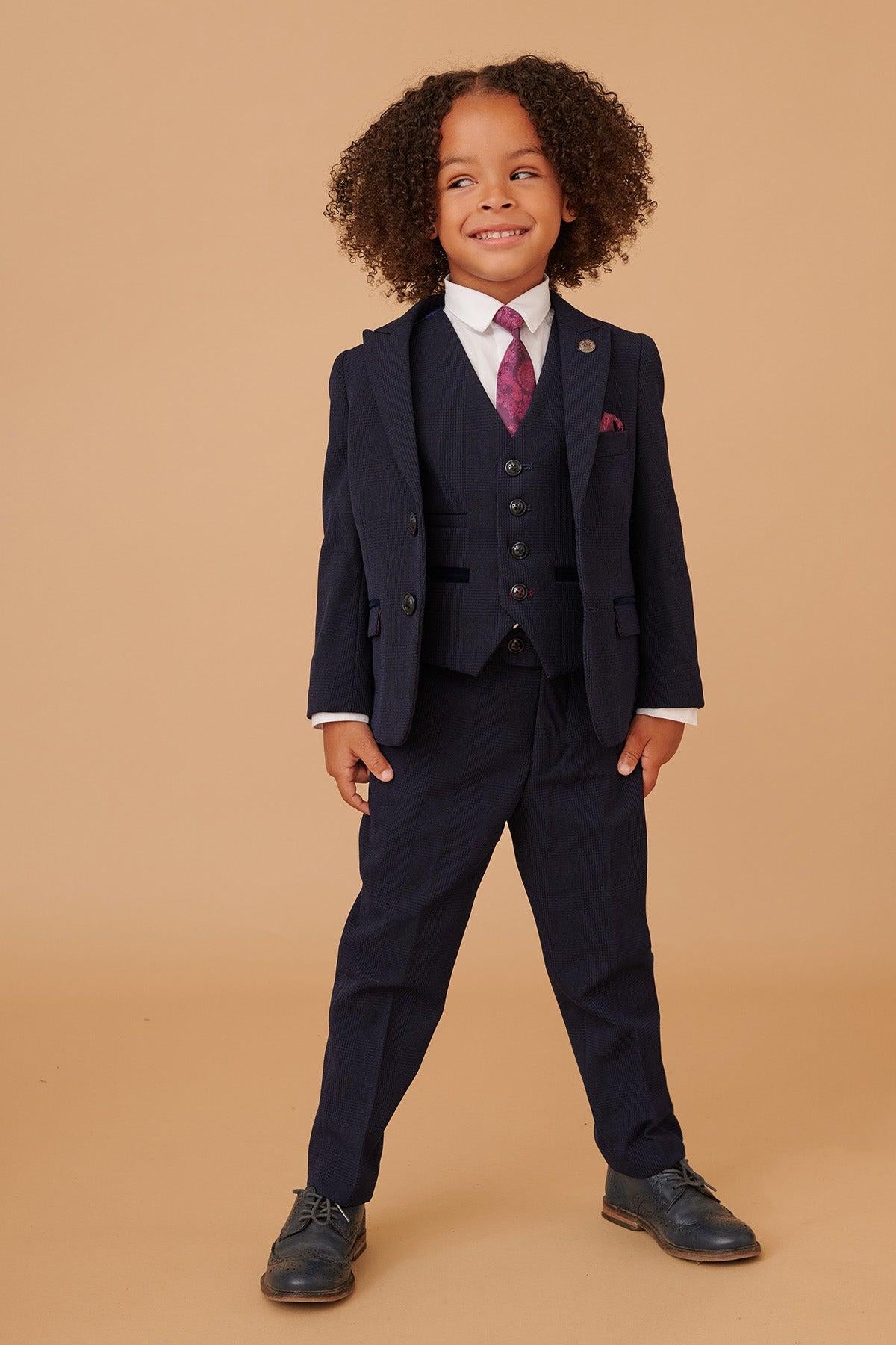 A young boy with curly hair smiles in the Marc Darcy BROMLEY Children's Navy Check Print Three Piece Suit, paired with a white shirt and patterned pink tie, posing against a tan background.
