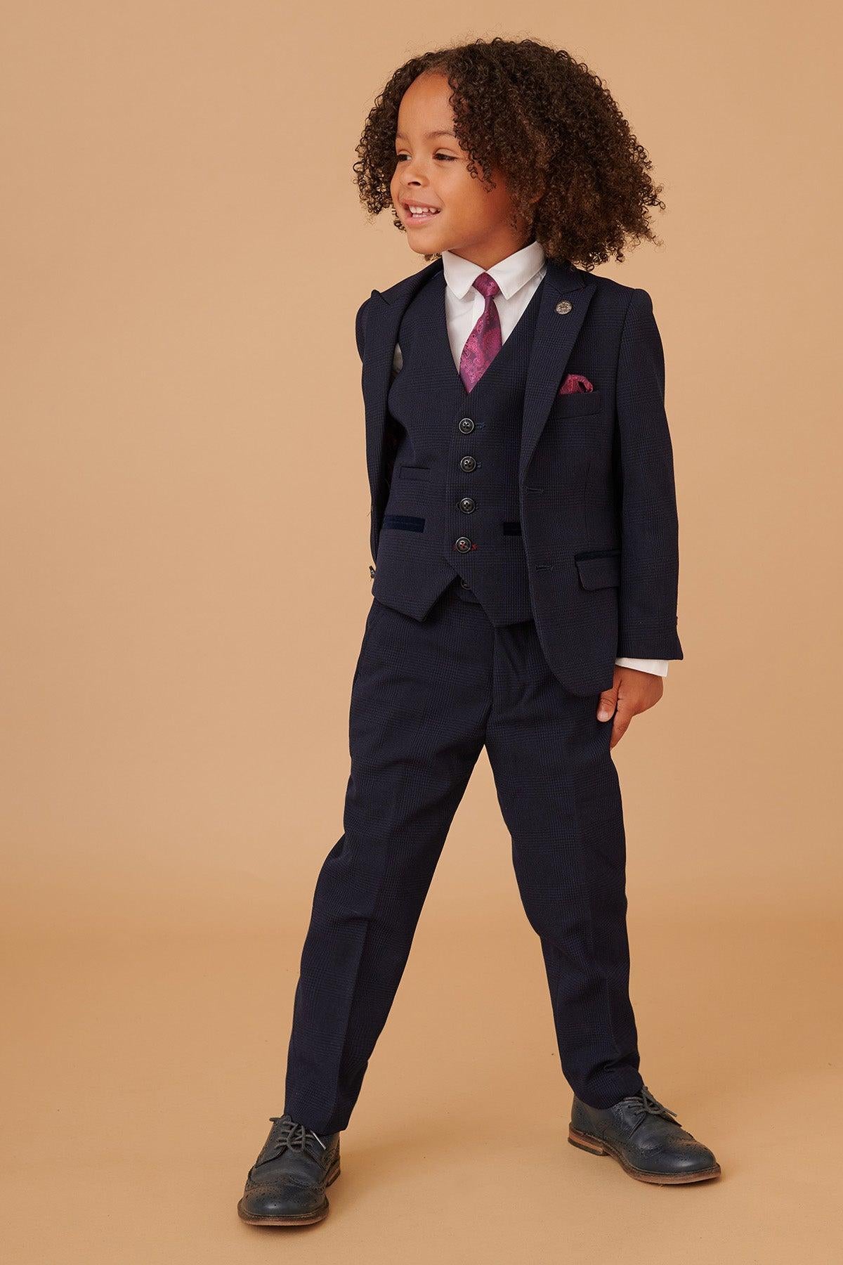 A young child with curly hair wears the Marc Darcy BROMLEY Children's Navy Check Print Three Piece Suit with an adjustable waist, white shirt, maroon tie, and black shoes, standing confidently against a beige background and smiling slightly.