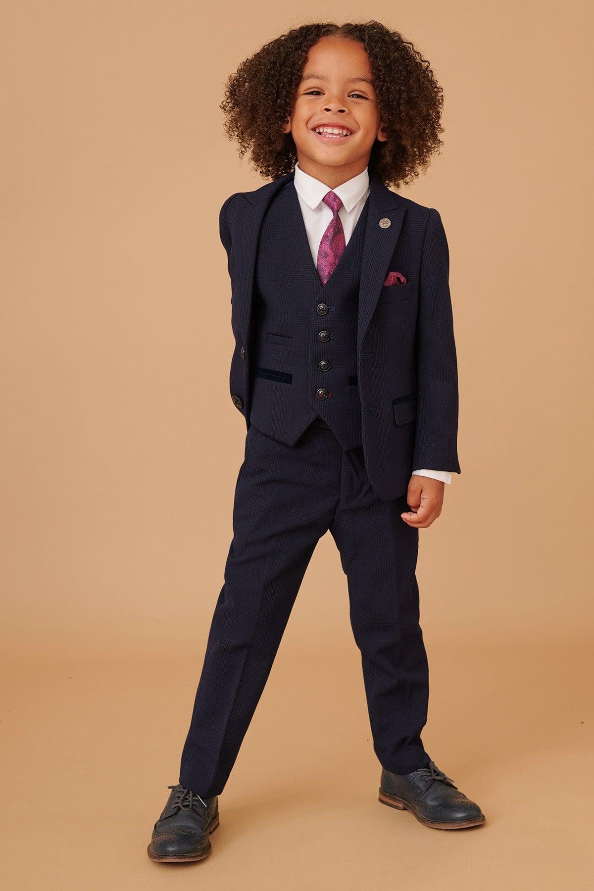 A young boy with curly hair smiles broadly, dressed in the Marc Darcy BROMLEY Children's Navy Check Print Three Piece Suit, paired with a patterned pink tie and white shirt—an outfit blending stylish flair with classic Prince of Wales check elegance.