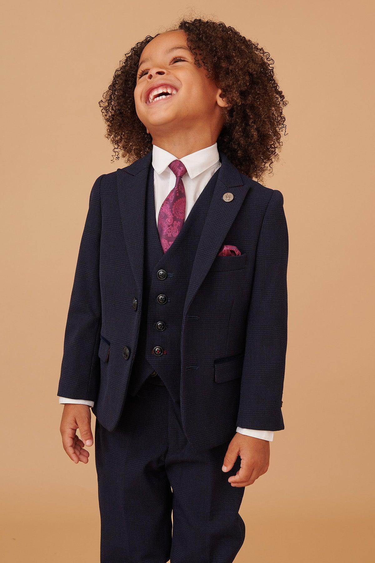 A young child in the Marc Darcy BROMLEY Children's Navy Check Print Three Piece Suit smiles and looks upward, dressed in a white shirt and purple patterned tie, against a plain beige background.