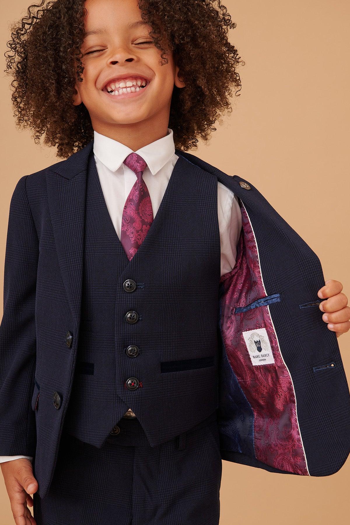 A young child with curly hair smiles widely, wearing the Marc Darcy BROMLEY Childrens Navy Check Print Three Piece Suit with an adjustable trouser waistband, white shirt, patterned purple tie, and showing off the suits colorful inner lining.