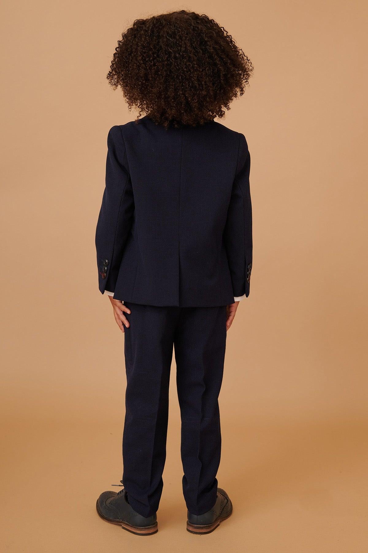 Back view of a child wearing the Marc Darcy BROMLEY children's navy check print three-piece suit with a white shirt and dress shoes against a plain tan background.
