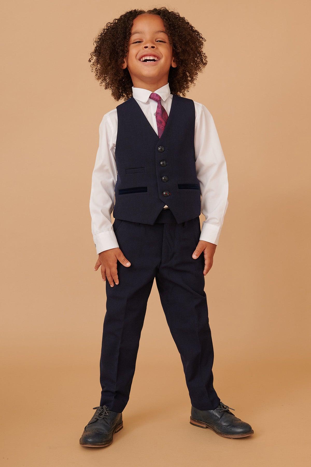 A young boy with curly hair smiles in the Marc Darcy BROMLEY Children's Navy Check Print Three Piece Suit, featuring a vest, adjustable waistband pants, white shirt, maroon tie, and black shoes against a plain beige background.