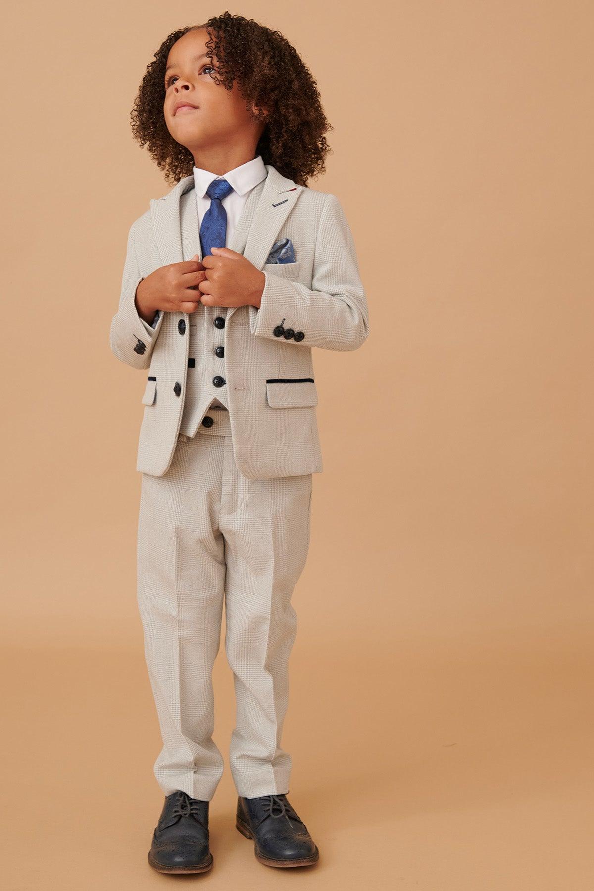 A young boy with curly brown hair looks up, wearing the BROMLEY Children's Stone Check Print Three Piece Suit by Marc Darcy, styled with a blue tie and pocket square.