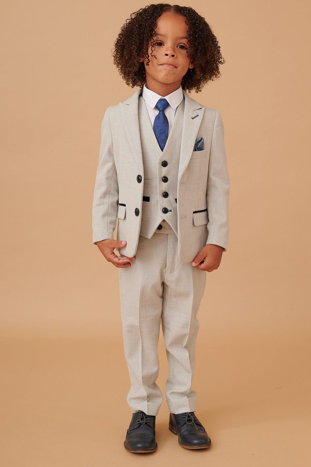 A young boy with curly hair models the Marc Darcy BROMLEY Childrens Stone Check Print Three Piece Suit, featuring an adjustable waistband, white shirt, blue tie and pocket square, and dark shoes, against a beige background.