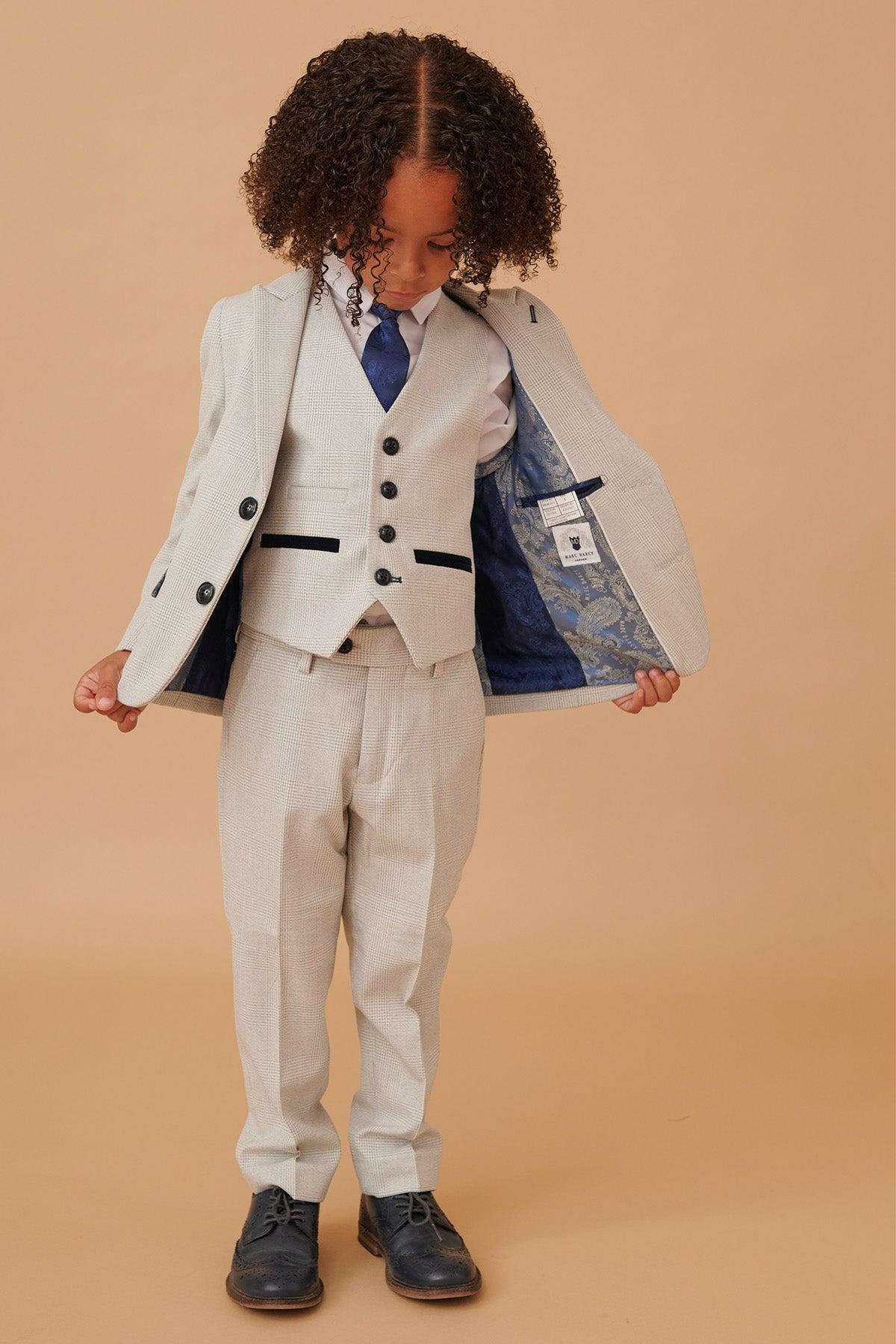 A young boy with curly hair models the Marc Darcy BROMLEY Children’s Stone Check Print Three Piece Suit, styled with a blue tie and lining.
