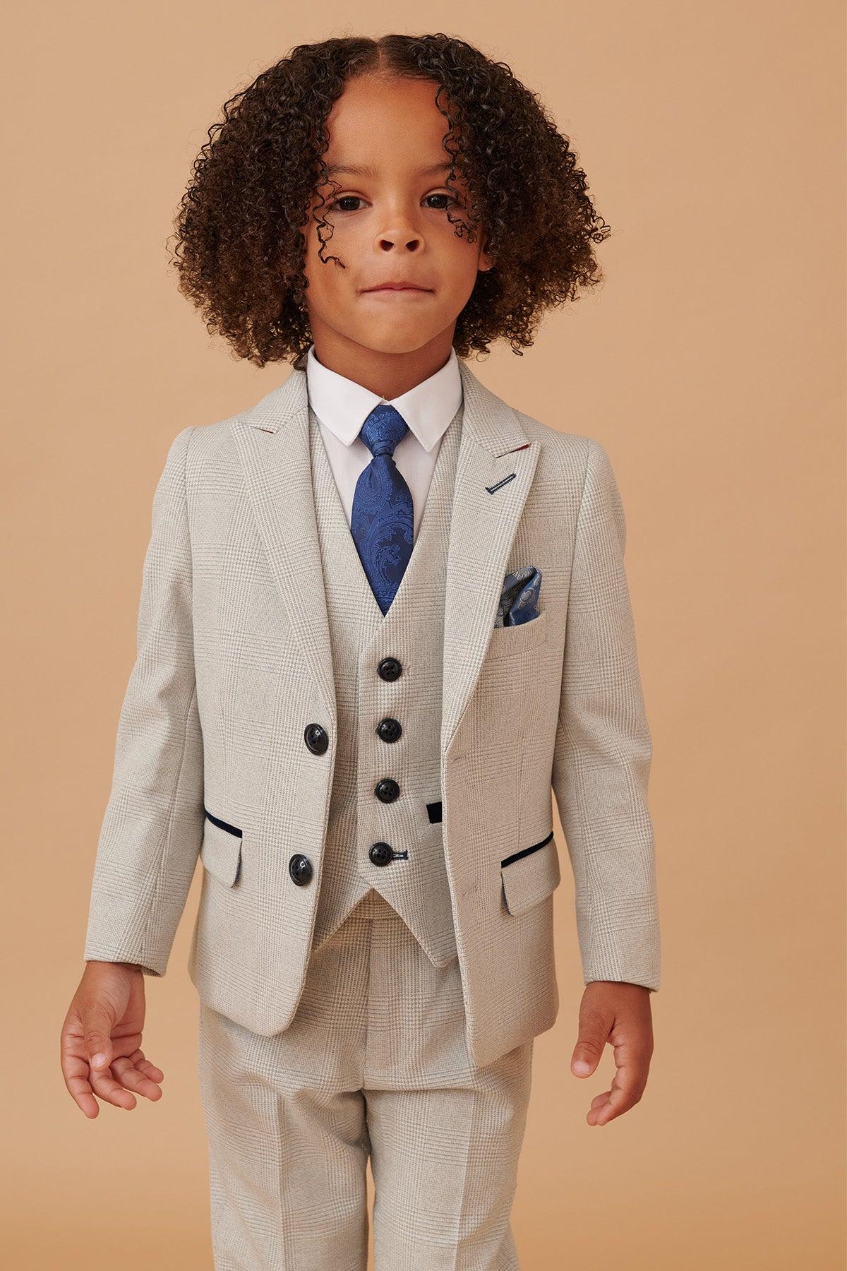 A young child with curly hair wears the Marc Darcy BROMLEY Children's Stone Check Print Three Piece Suit, paired with a white shirt and blue tie, standing against a plain beige background.