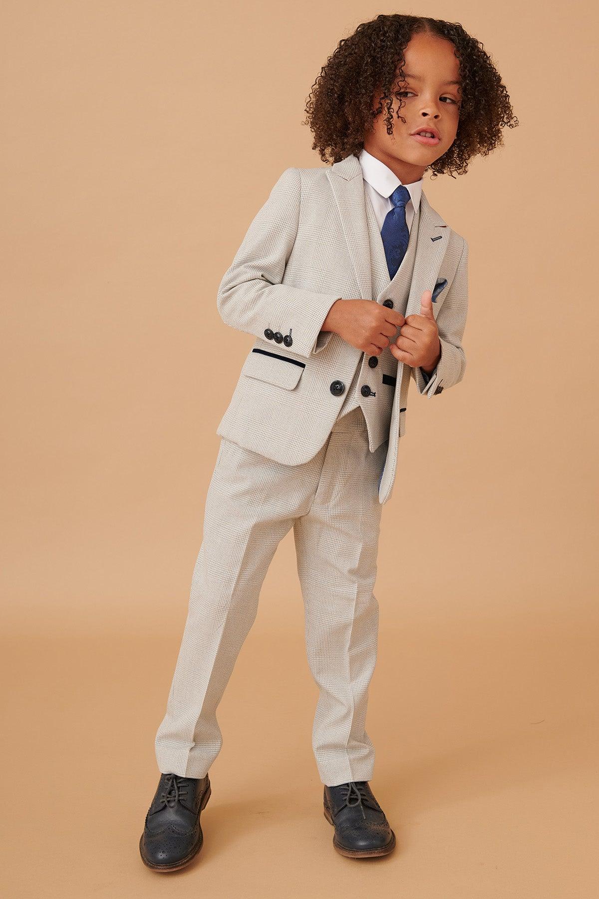 A young boy poses in the Marc Darcy BROMLEY Children’s Stone Check Print Three Piece Suit with a white shirt, blue tie, and dark brogue shoes, set against a tan background. Matching adult suit also available.