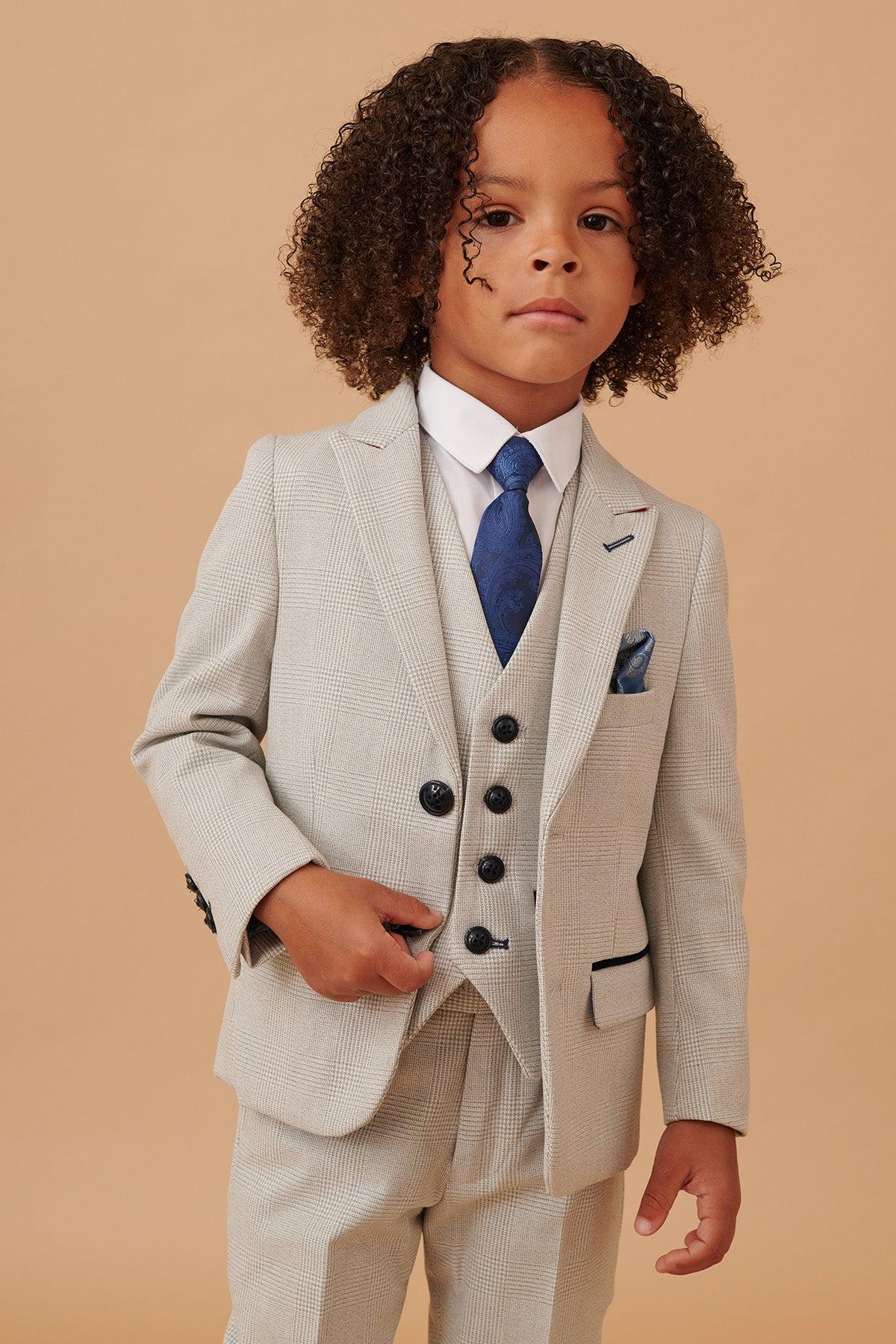 A young boy with curly hair wears the Marc Darcy BROMLEY Children's Stone Check Print Three Piece Suit, paired with a white shirt, blue patterned tie, and blue pocket square.