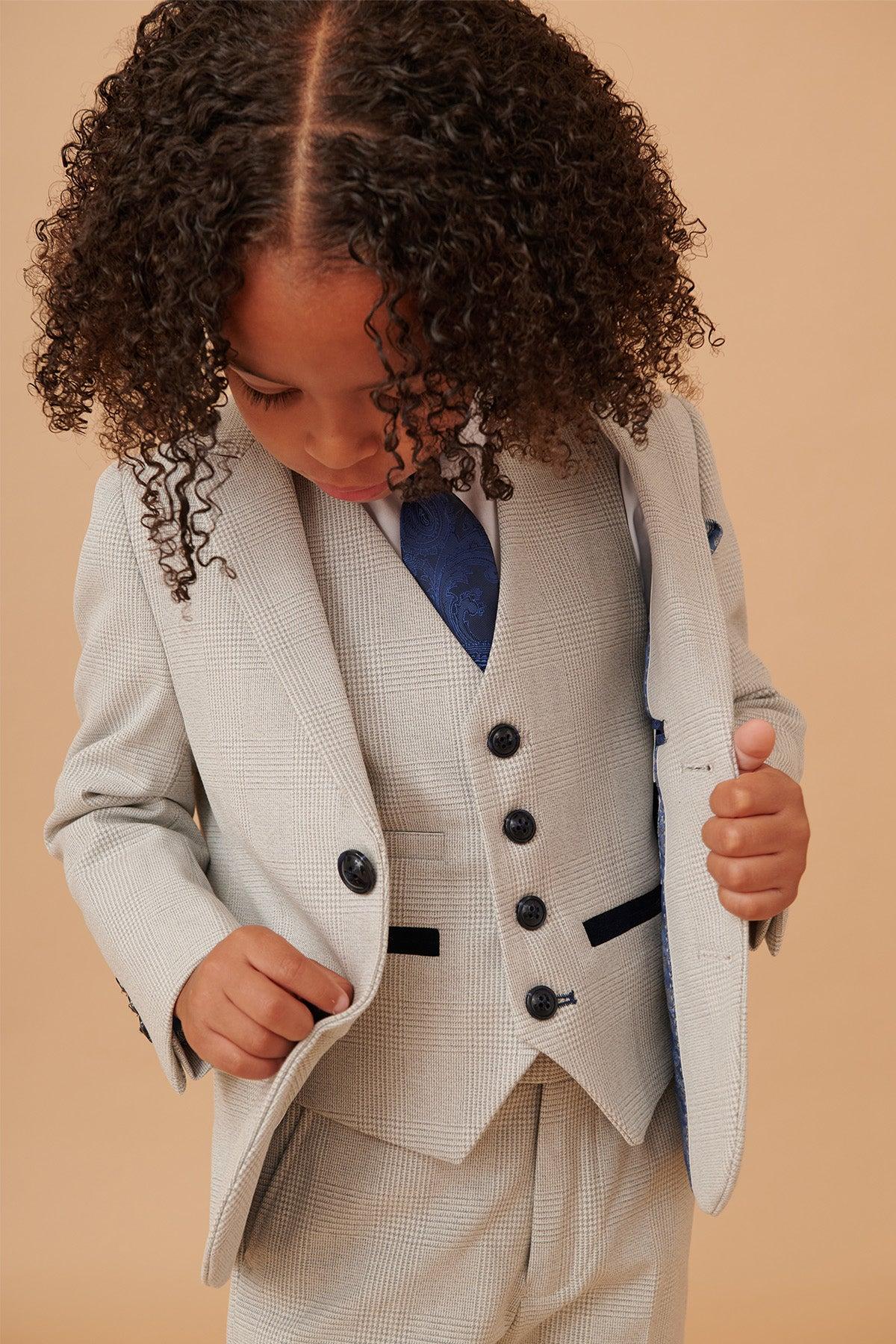 A young child in the Marc Darcy BROMLEY Childrens Stone Check Print Three Piece Suit looks down while adjusting the jacket, wearing a blue patterned tie and white shirt against a neutral beige background.