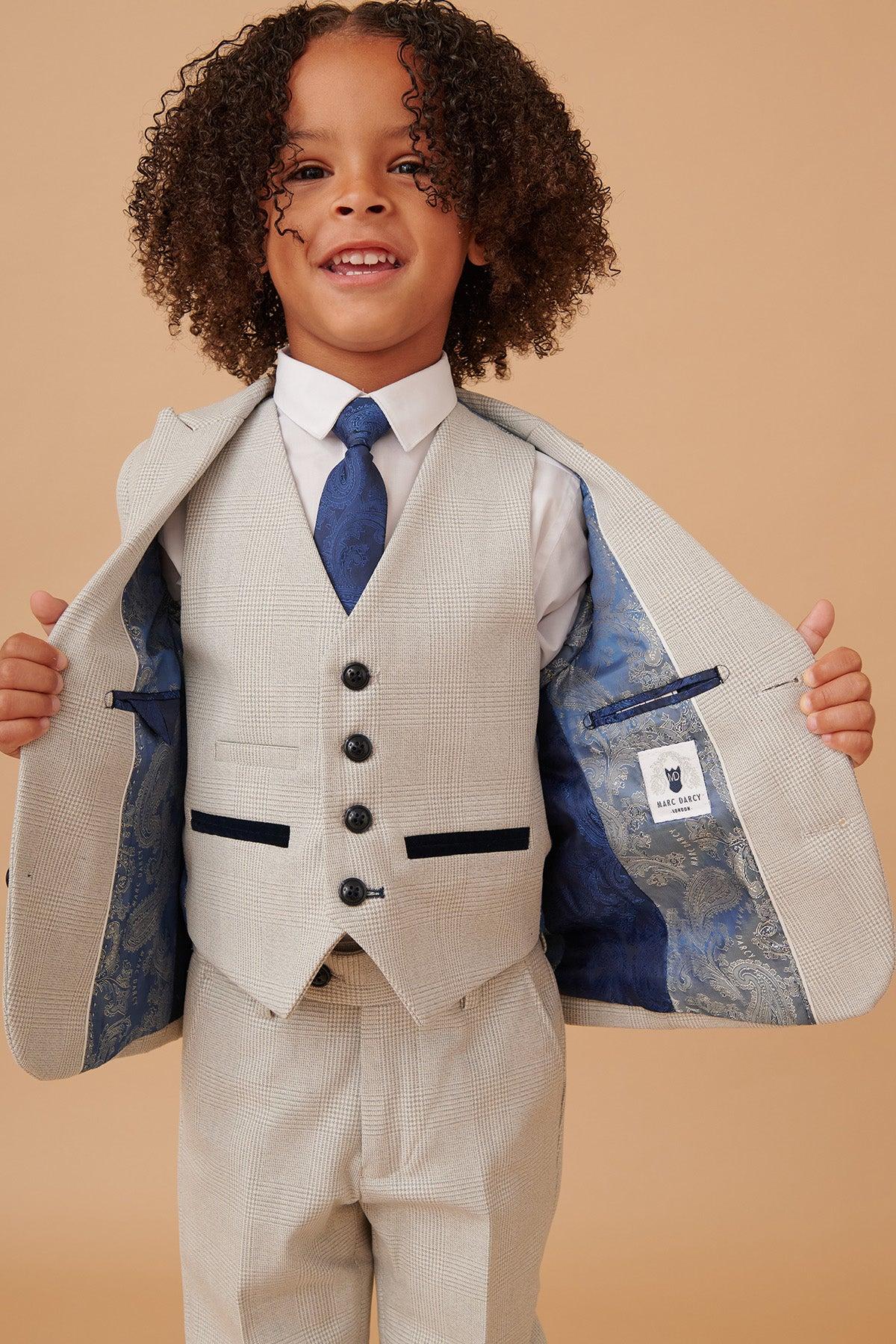 A young boy wears the BROMLEY Children's Stone Check Print Three Piece Suit by Marc Darcy with a white shirt and blue patterned tie, opening his jacket to reveal its navy paisley lining.