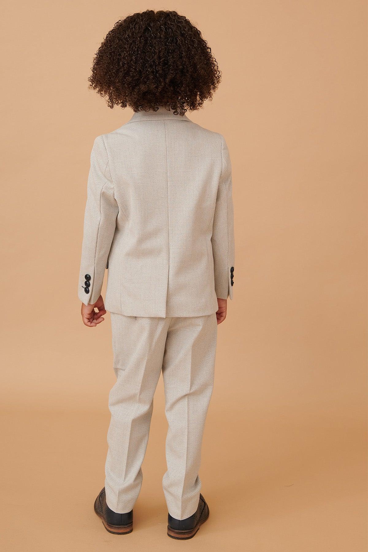 A child with curly hair wears the Marc Darcy BROMLEY Childrens Stone Check Print Three Piece Suit, featuring an adjustable waistband, and black shoes, standing with their back to the camera against a plain beige background.
