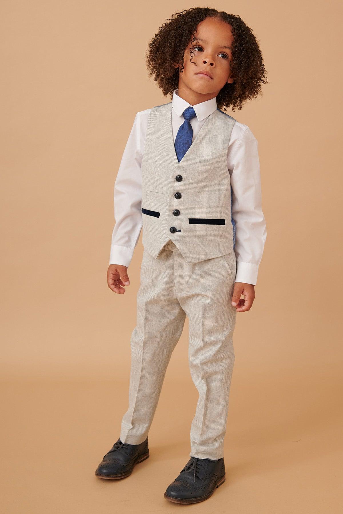 A young child with curly hair wears the Marc Darcy BROMLEY Childrens Stone Check Print Three Piece Suit with a white shirt, blue tie, and black shoes, standing against a plain beige background.