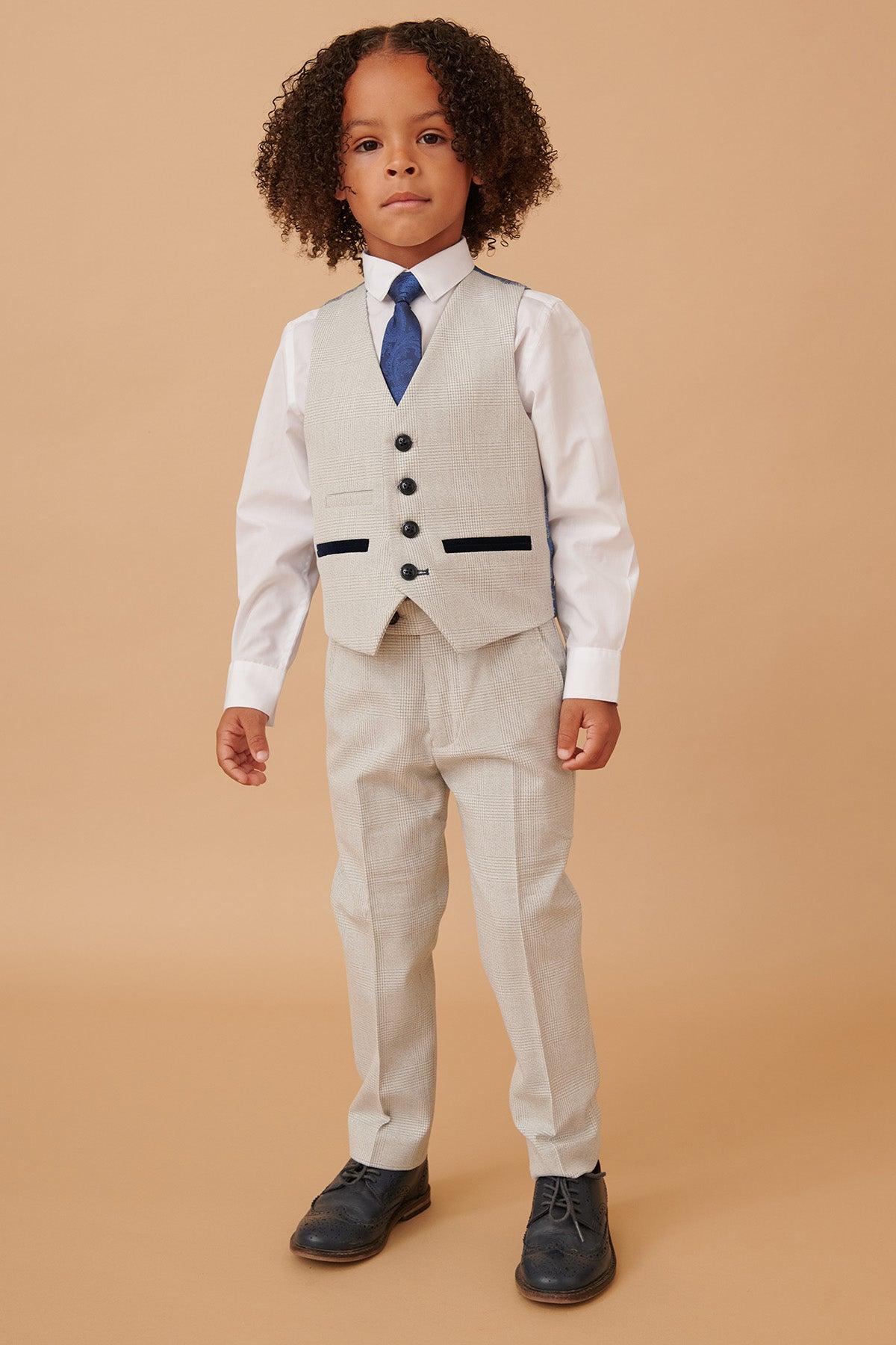 A young boy wears the Marc Darcy BROMLEY Children's Stone Check Print Three Piece Suit with a white shirt, blue tie, and black shoes, standing against a plain beige background.