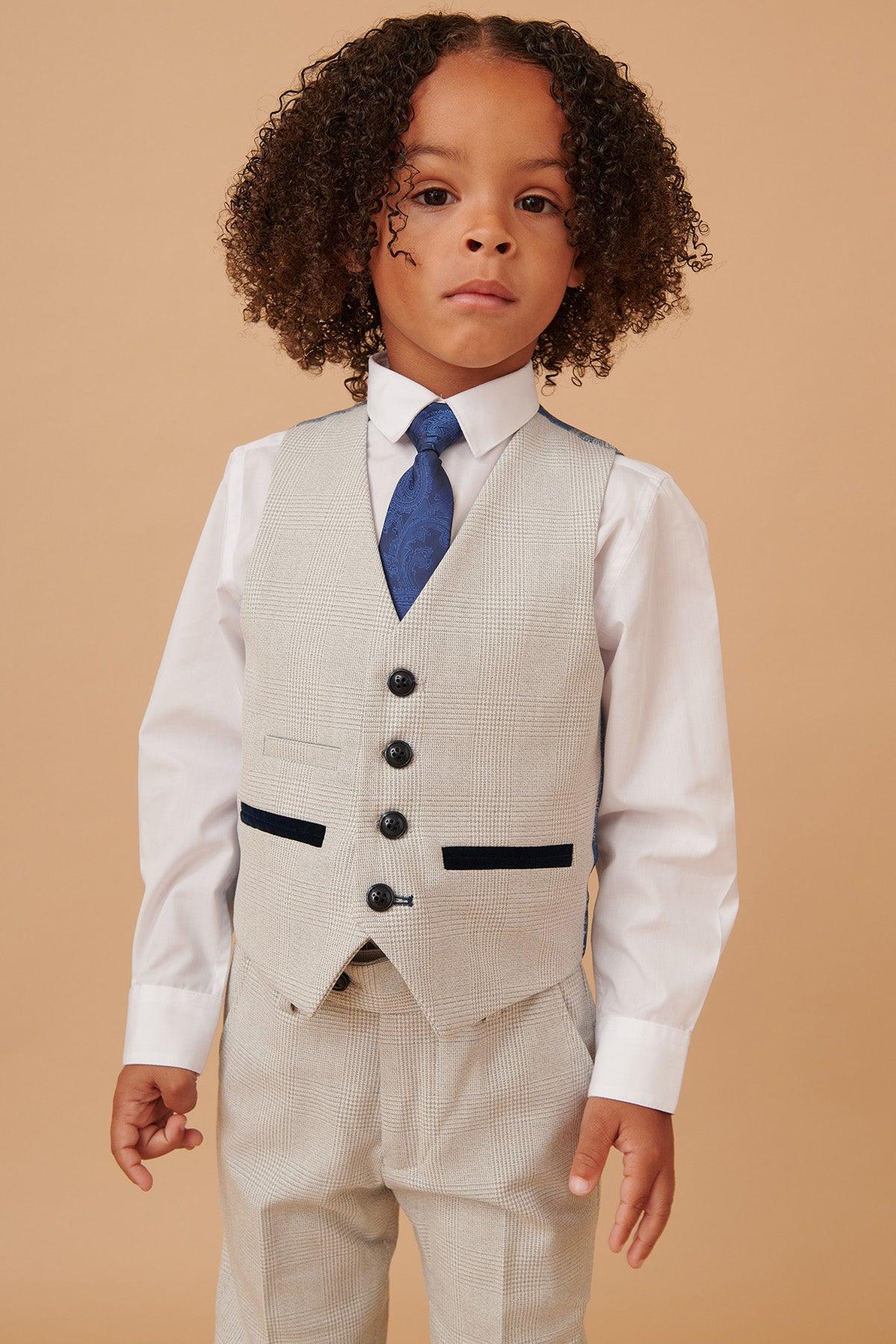 A young child with curly hair wears the Marc Darcy BROMLEY Children's Stone Check Print Three Piece Suit, featuring a matching vest and trousers, along with a white shirt and blue patterned tie, standing against a beige background.