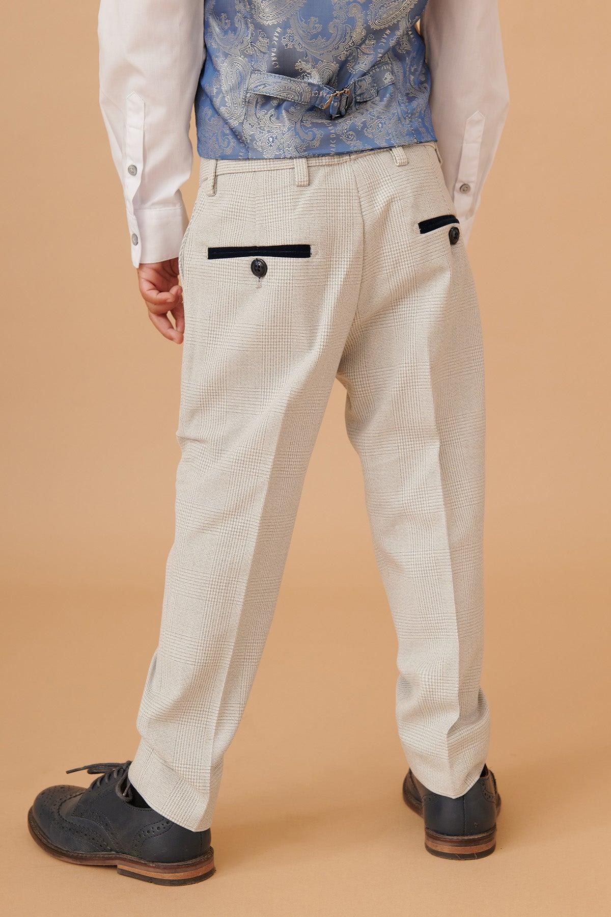 A child faces away from the camera in the Marc Darcy BROMLEY Stone Check Three Piece Suit, featuring light check pants with black-trimmed pockets and buttons, a white shirt, a blue patterned vest, black dress shoes, and an adjustable waistband for comfort.