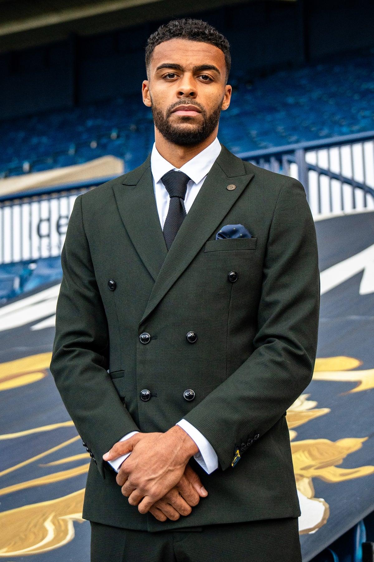A young Black man stands with his arms crossed in front of a stadium, wearing the Marc Darcy BROMLEY Olive Green Double Breasted Check Blazer, paired with a white shirt and black tie.