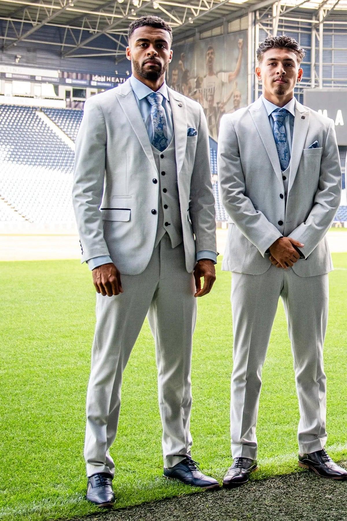 Two men wear Marc Darcy's The WBA Collection - BROMLEY Stone Suits and blue patterned ties, standing on a stadium field. Both have short hair and face the camera, with stadium seats and a large billboard behind them.