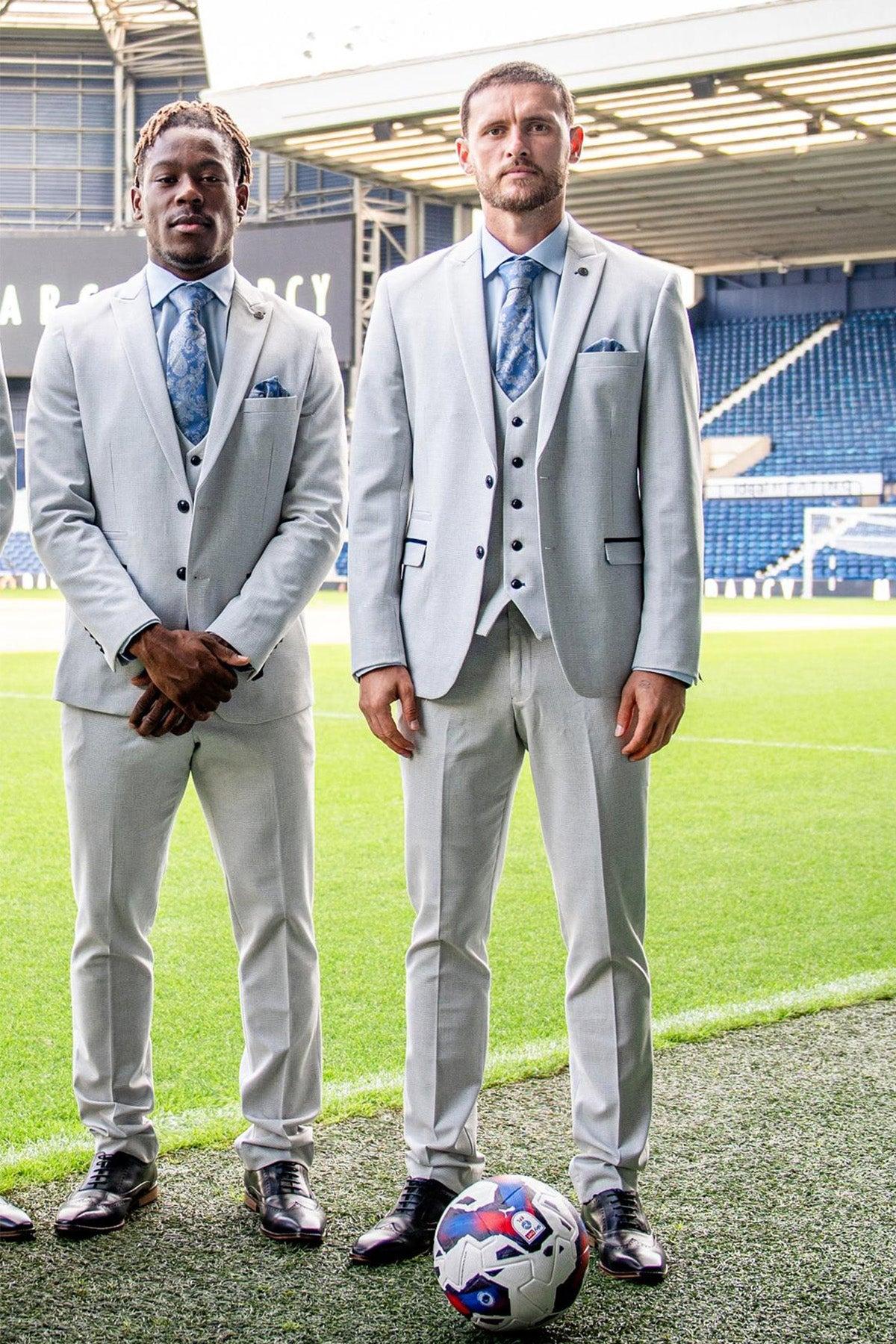 Two men stand on a soccer field in Marc Darcy's The WBA Collection - BROMLEY Stone three-piece suits, with blue ties and vests. One rests his hands in front; the other stands with a foot on a soccer ball. Empty stadium seats are behind them.