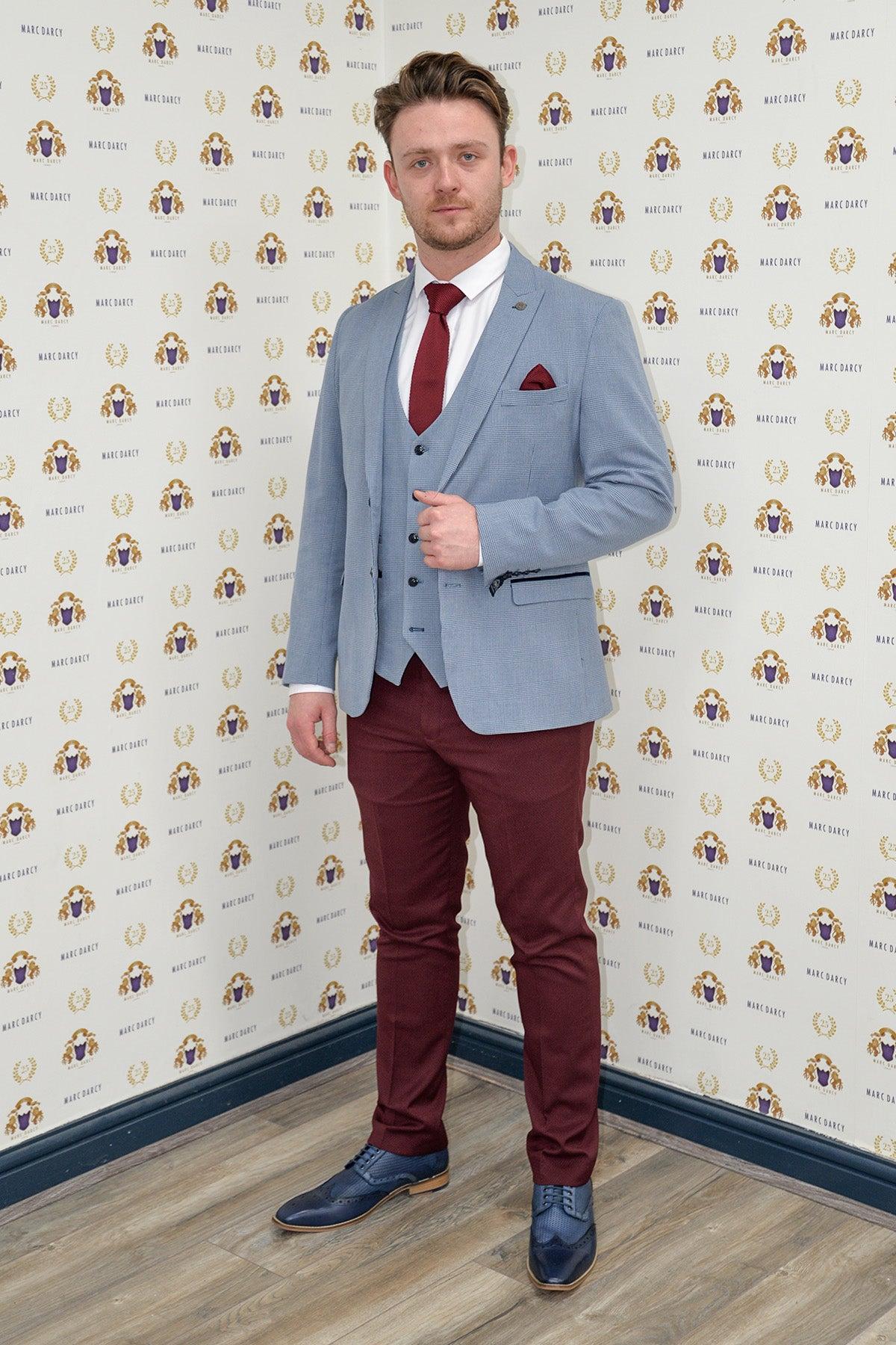 Calum Lill as Joel Deering wears the Marc Darcy BROMLEY Sky Blue Blazer with wine chinos on a wood floor in front of a patterned wall with repeated logos.