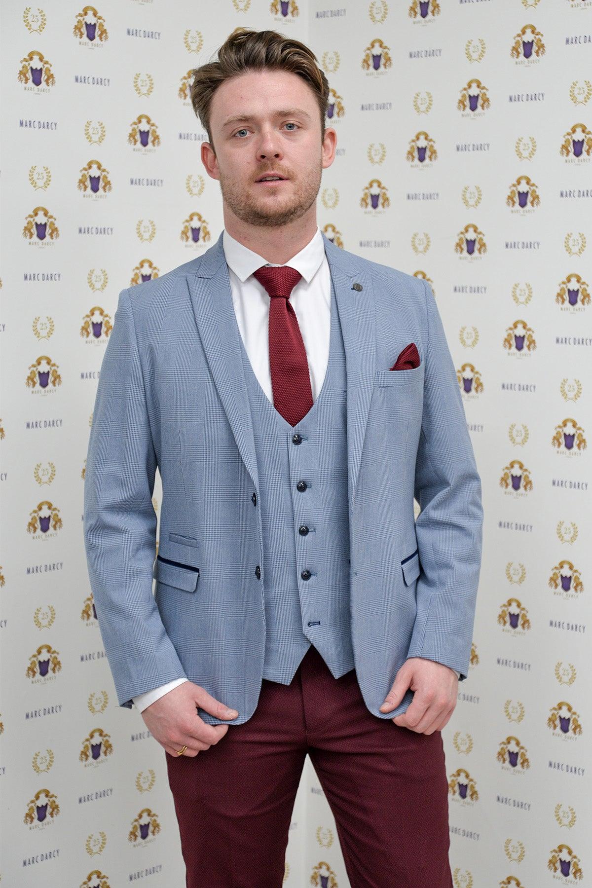 Calum Lill as Coronation Street’s Joel Deering wears the Marc Darcy BROMLEY Sky Blue Blazer with wine chinos, standing before a gold and blue branded backdrop.