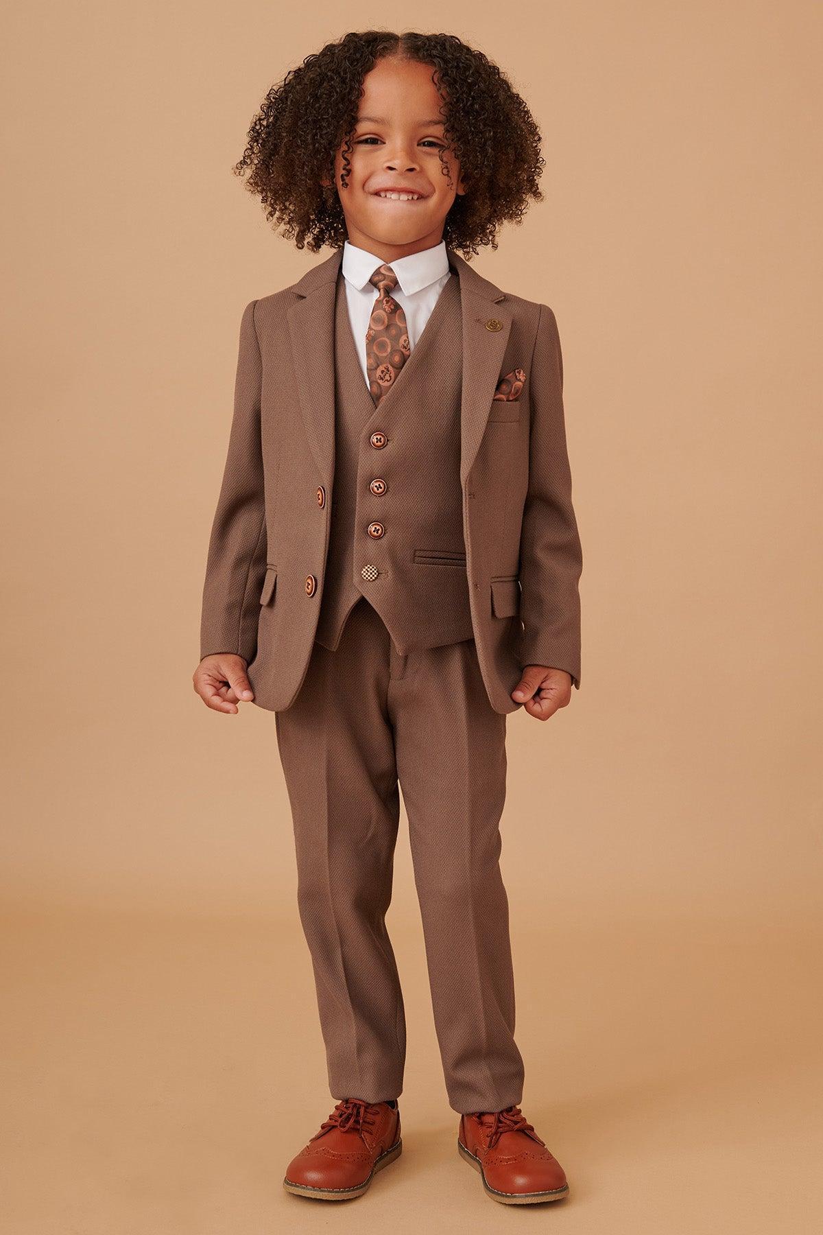 A young child with curly hair beams in the Marc Darcy HM5 - Childrens Tan Tailored Three Piece Suit, paired with a white shirt, patterned tie, and brown dress shoes—all set against a beige backdrop.