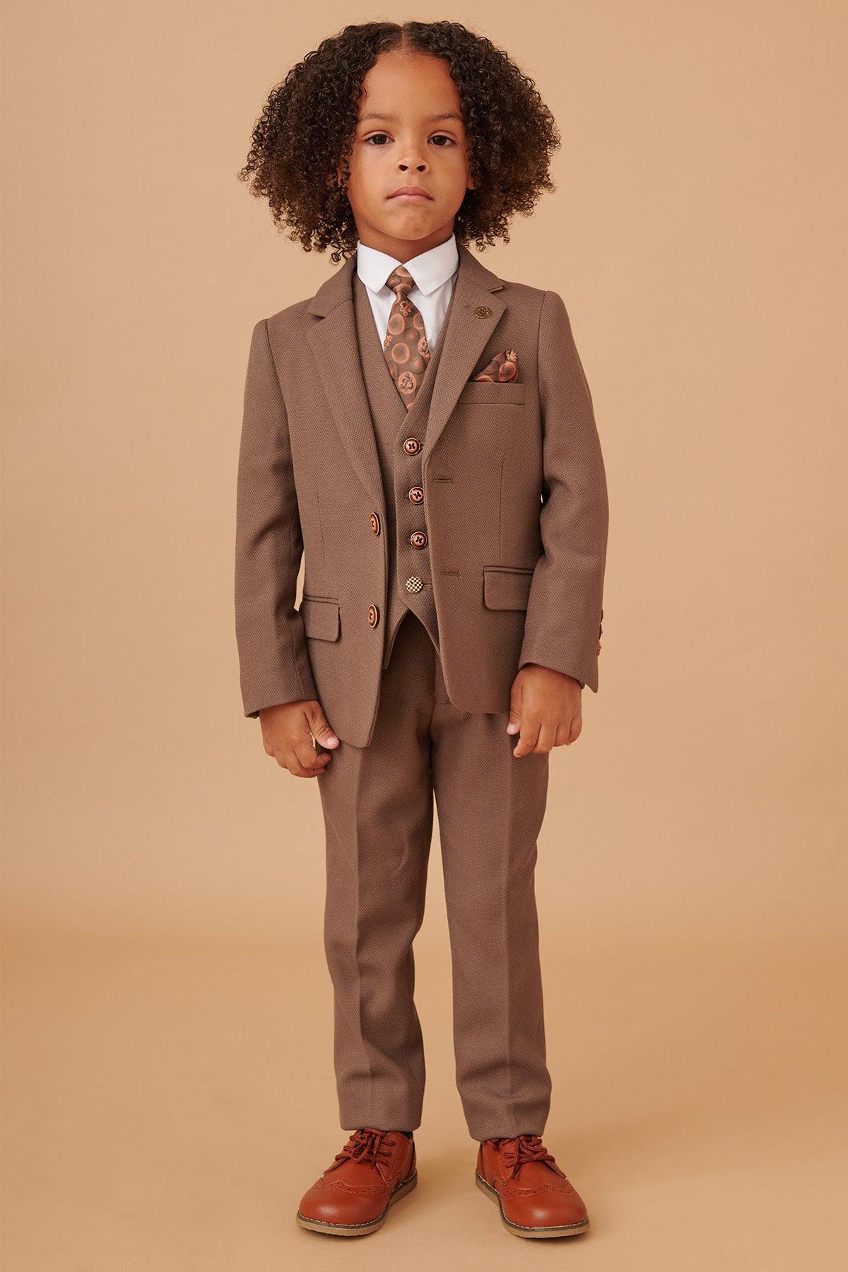 A young boy with curly hair models the Marc Darcy HM5 - Children's Tan Tailored Three Piece Suit, complete with a white shirt, patterned tie, pocket square, and brown lace-up shoes—ideal for weddings and formal occasions.