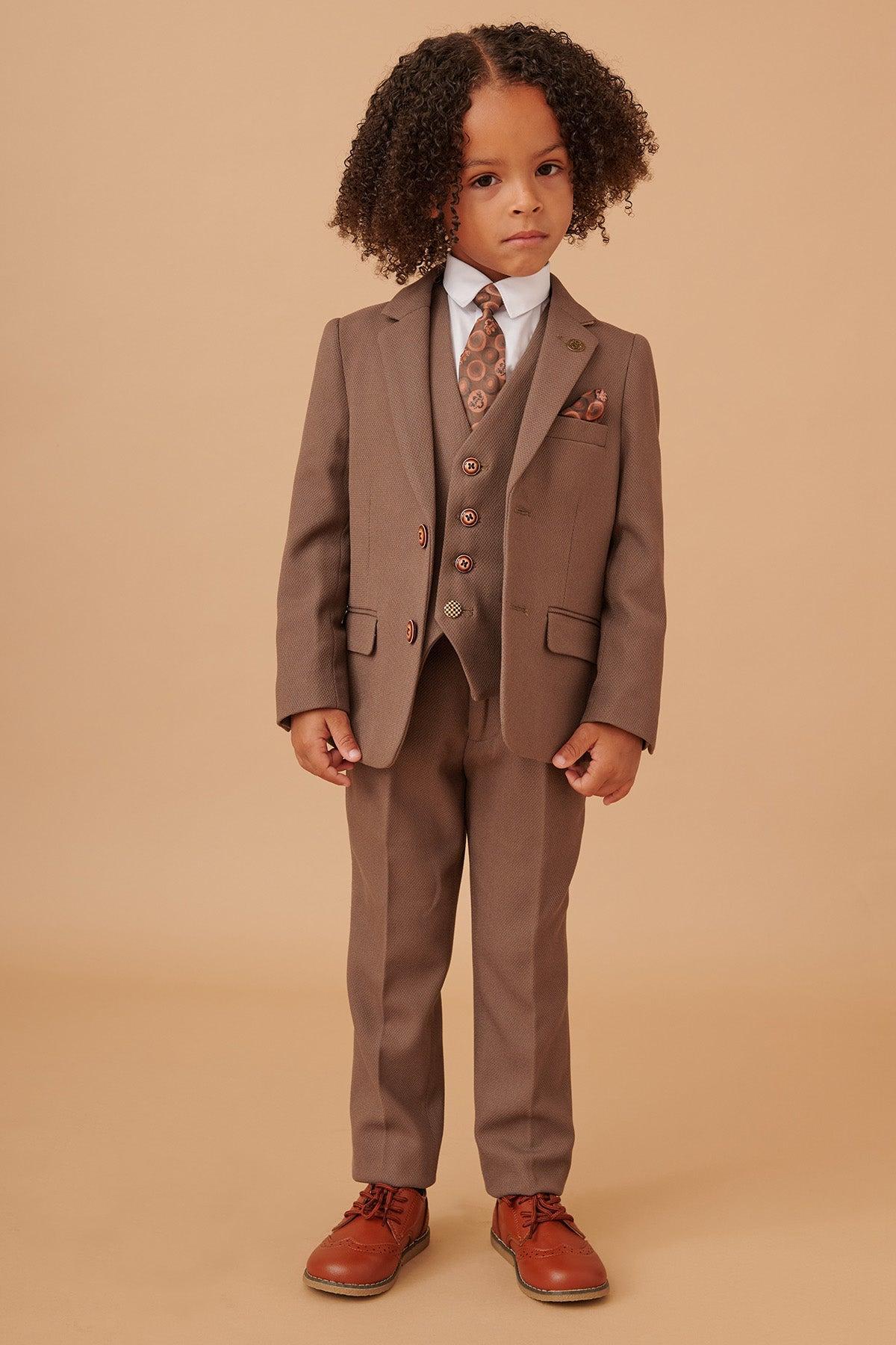 A young boy with curly hair models the Marc Darcy HM5 - Children's Tan Tailored Three Piece Suit, paired with a white shirt, patterned tie, and brown dress shoes—ideal formalwear for weddings.