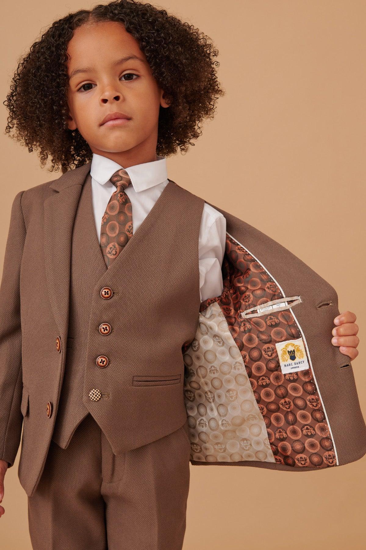 A young boy wears the Marc Darcy HM5 Children's Tan Tailored Three Piece Suit with a white shirt and patterned tie. The open jacket reveals a colorful lining—ideal as a formal kids' suit for weddings.