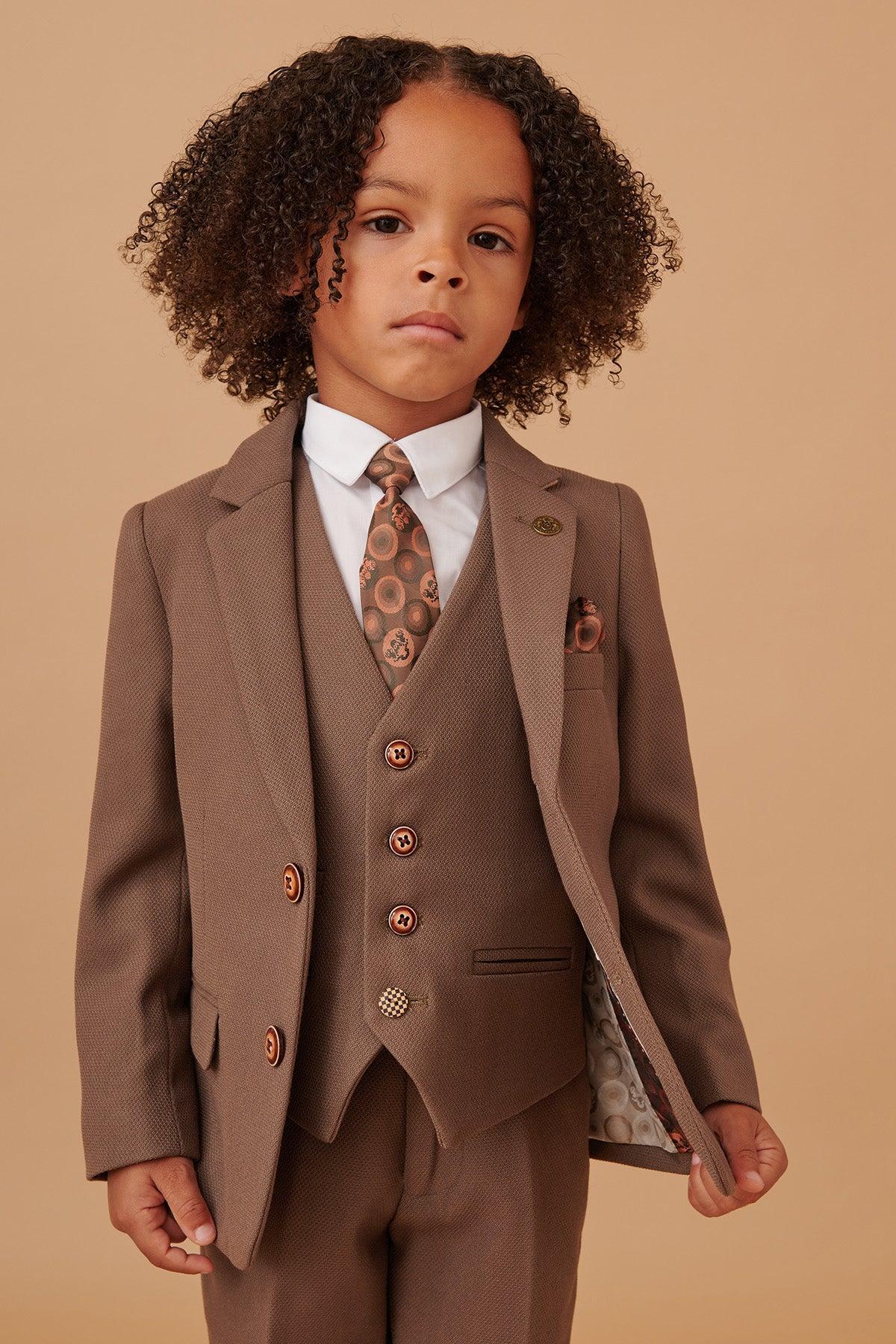 A young child with curly hair wears the Marc Darcy HM5 - Childrens Tan Tailored Three Piece Suit, featuring a brown suit, matching vest, white shirt, and patterned tie. Perfect formal wear for kids.