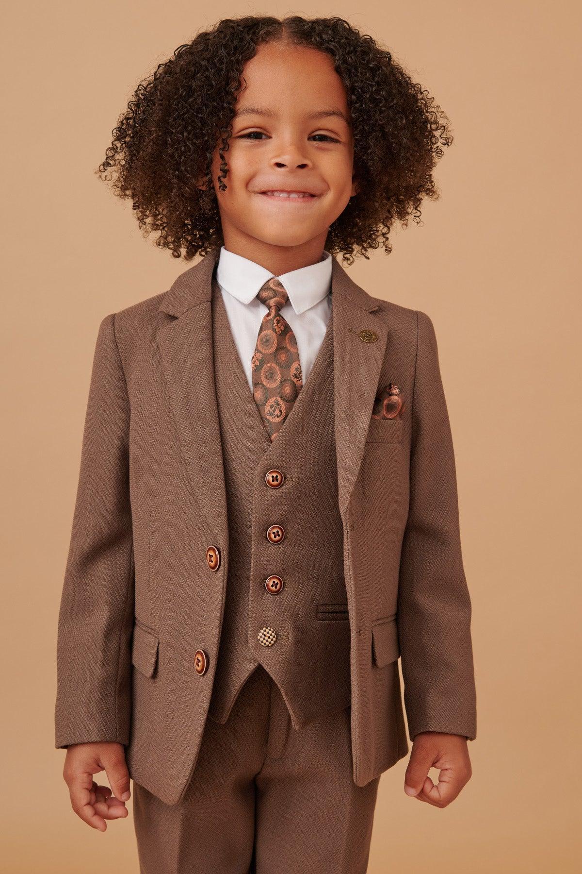 A young child with curly hair smiles while wearing the Marc Darcy HM5 Children's Tan Tailored Three Piece Suit with matching vest, white shirt, and patterned tie, standing in front of a beige background.