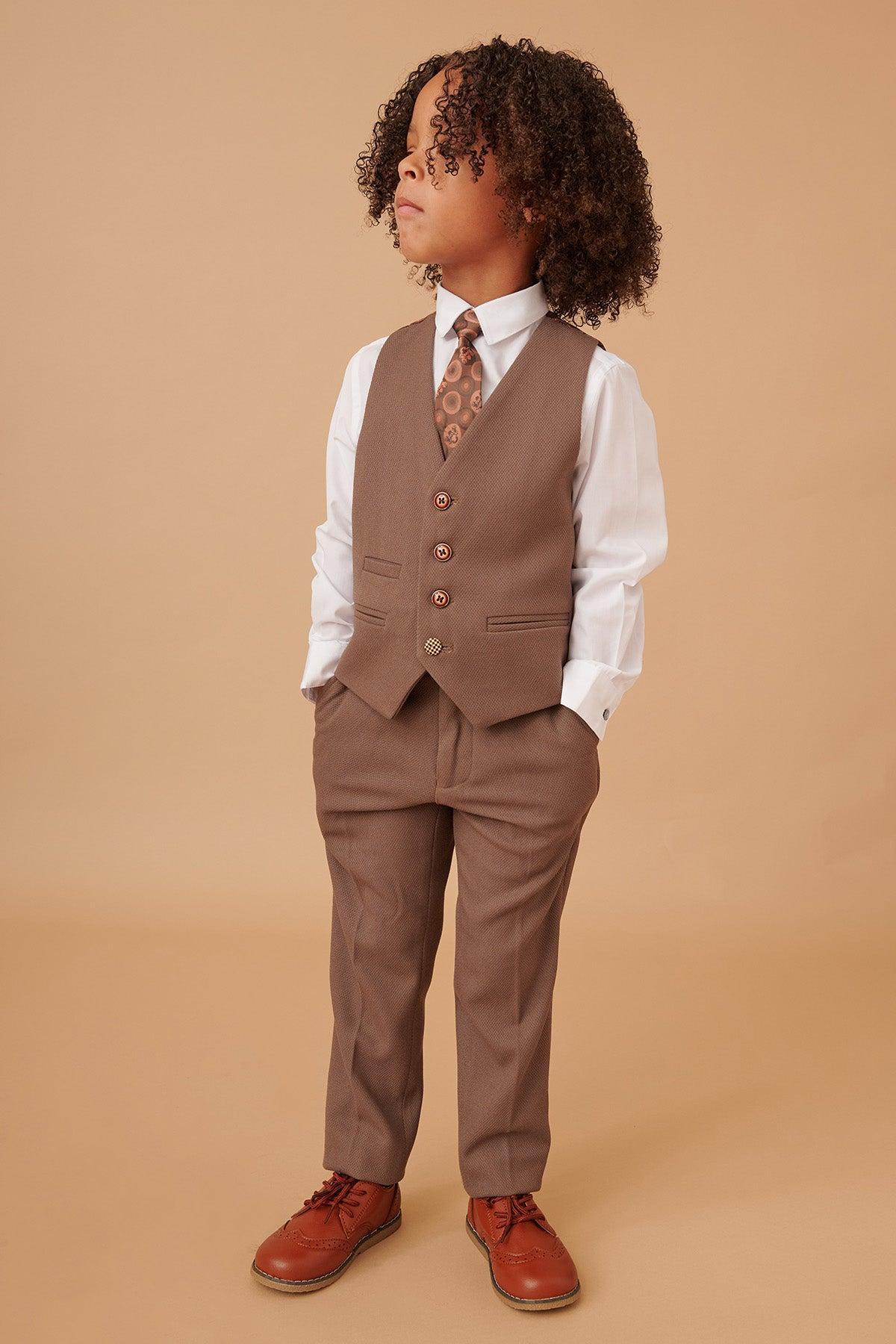 A young child with curly hair stands with hands in pockets, wearing the Marc Darcy HM5 - Children's Tan Tailored Three Piece Suit—featuring a vest, matching pants, white shirt, patterned tie, and brown shoes—set against a plain beige background.