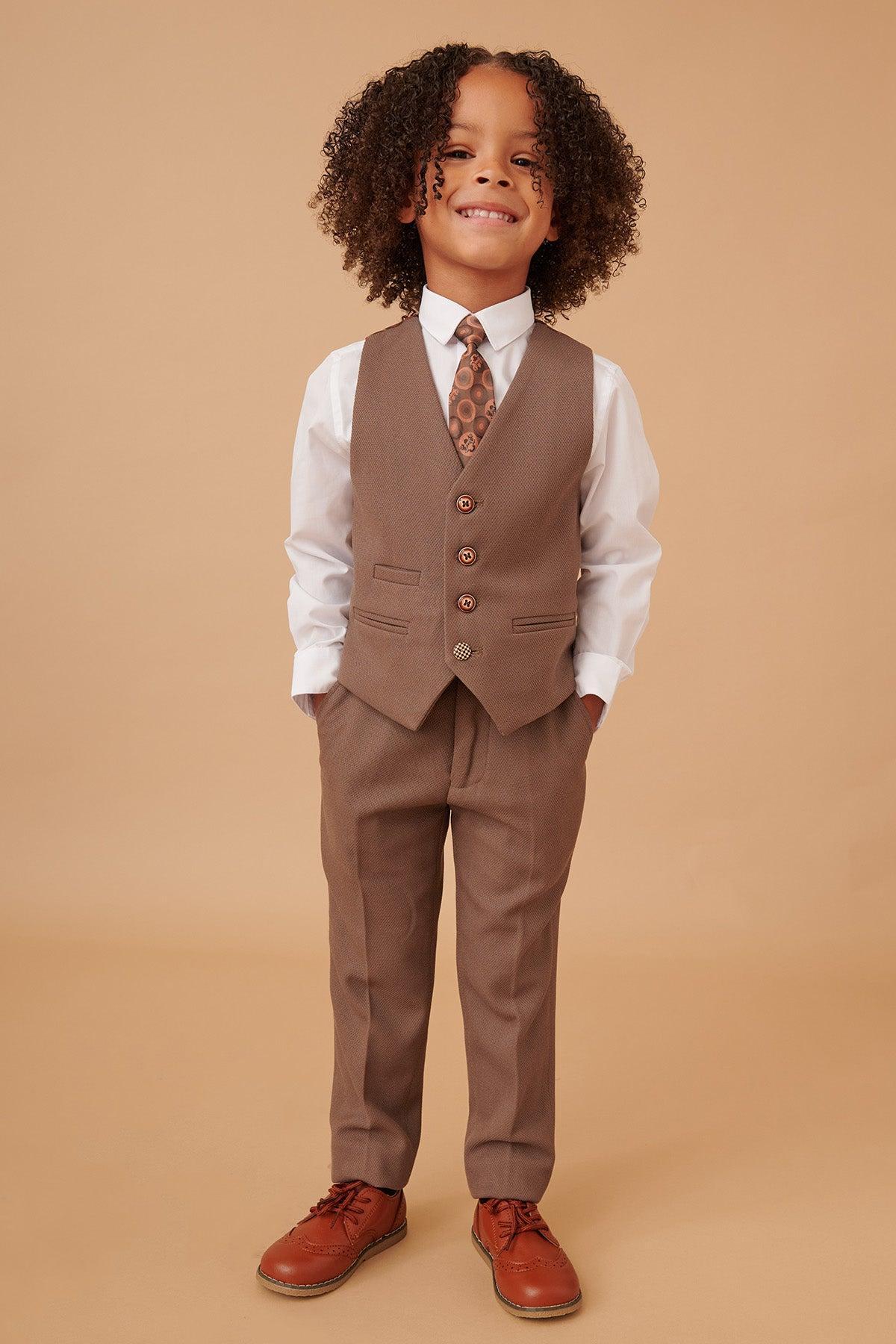 A young child with curly hair smiles in the Marc Darcy HM5 - Childrens Tan Tailored Three Piece Suit, featuring a brown vest, matching pants, white shirt, patterned tie, and brown dress shoes against a beige background.