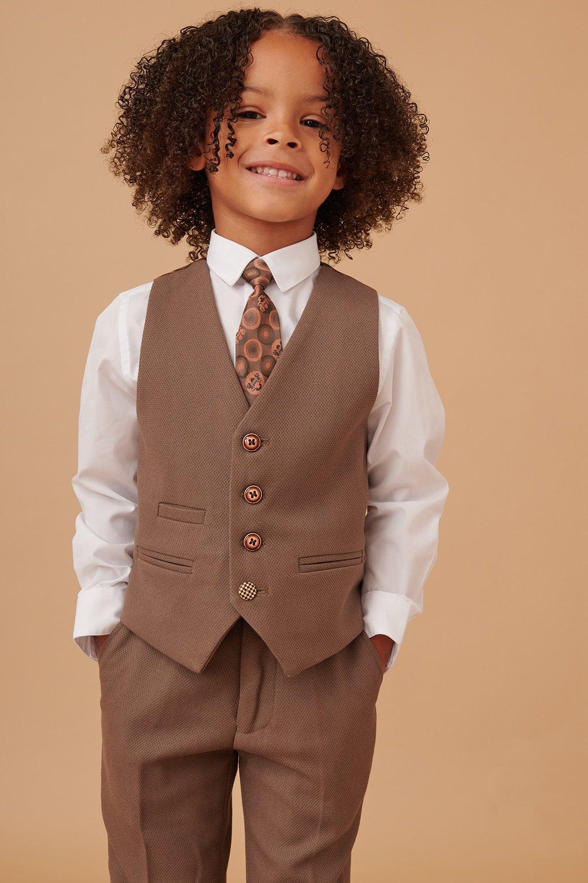 A young child with curly hair smiles, wearing the Marc Darcy HM5 - Childrens Tan Tailored Three Piece Suit—crafted for formal occasions—while standing against a beige backdrop with hands in pockets.