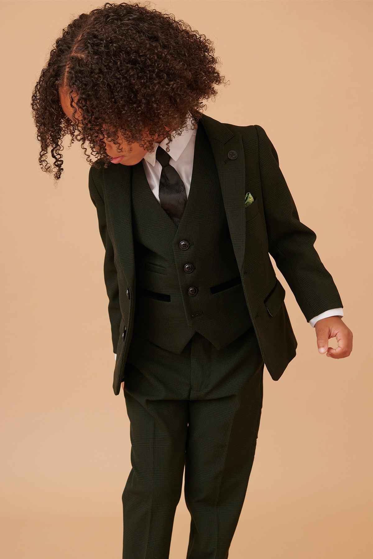 A young child with curly hair looks down while wearing the Marc Darcy BROMLEY Children's Olive Green Check Print Three Piece Suit, standing against a plain tan background.