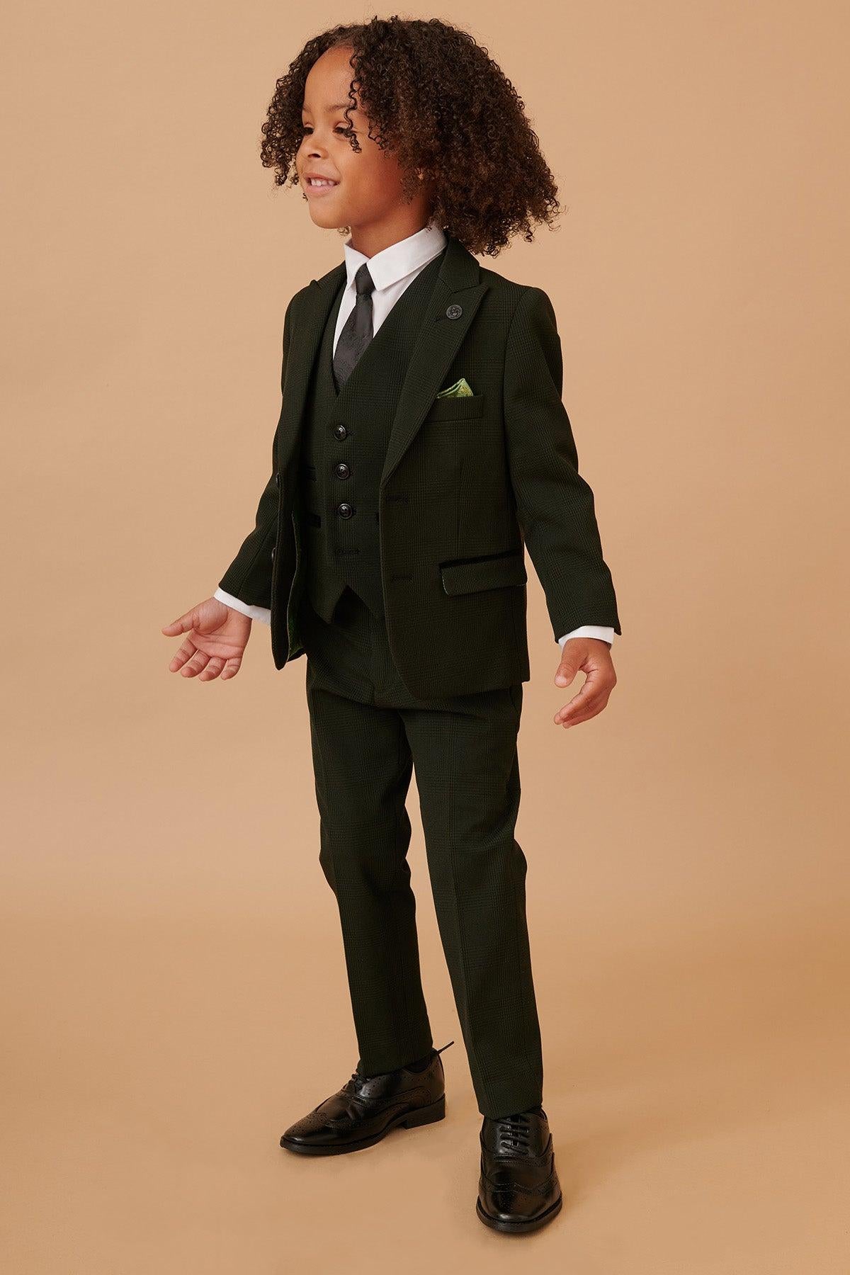 A young child with curly hair smiles while wearing the Marc Darcy BROMLEY Childrens Olive Green Check Print Three Piece Suit, white shirt, black tie, and black shoes, standing against a beige background.