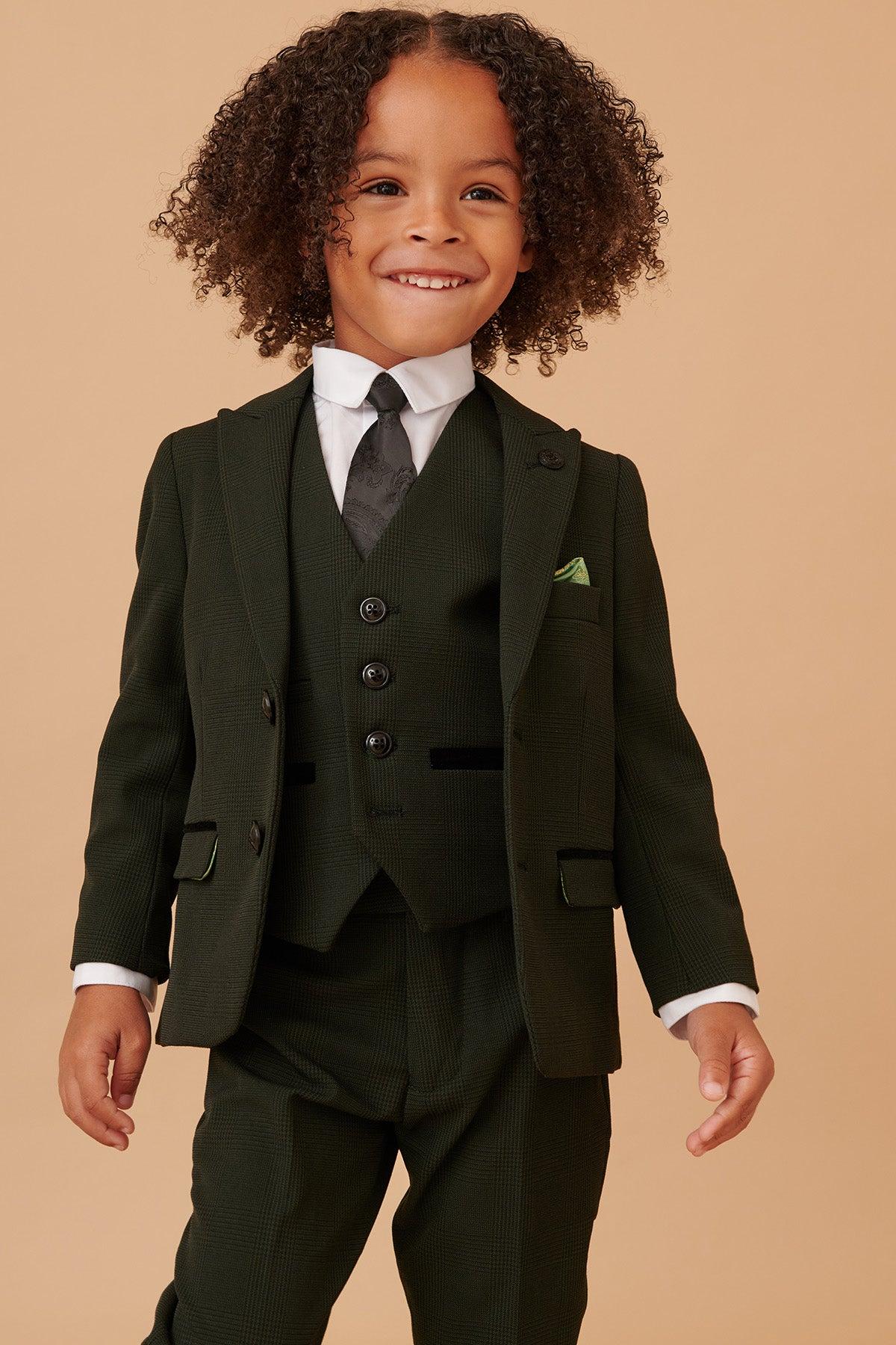 Smiling young child with curly hair wearing the Marc Darcy BROMLEY childrens olive green check print three piece suit, featuring a matching vest, white shirt, and black tie, standing against a plain beige background.