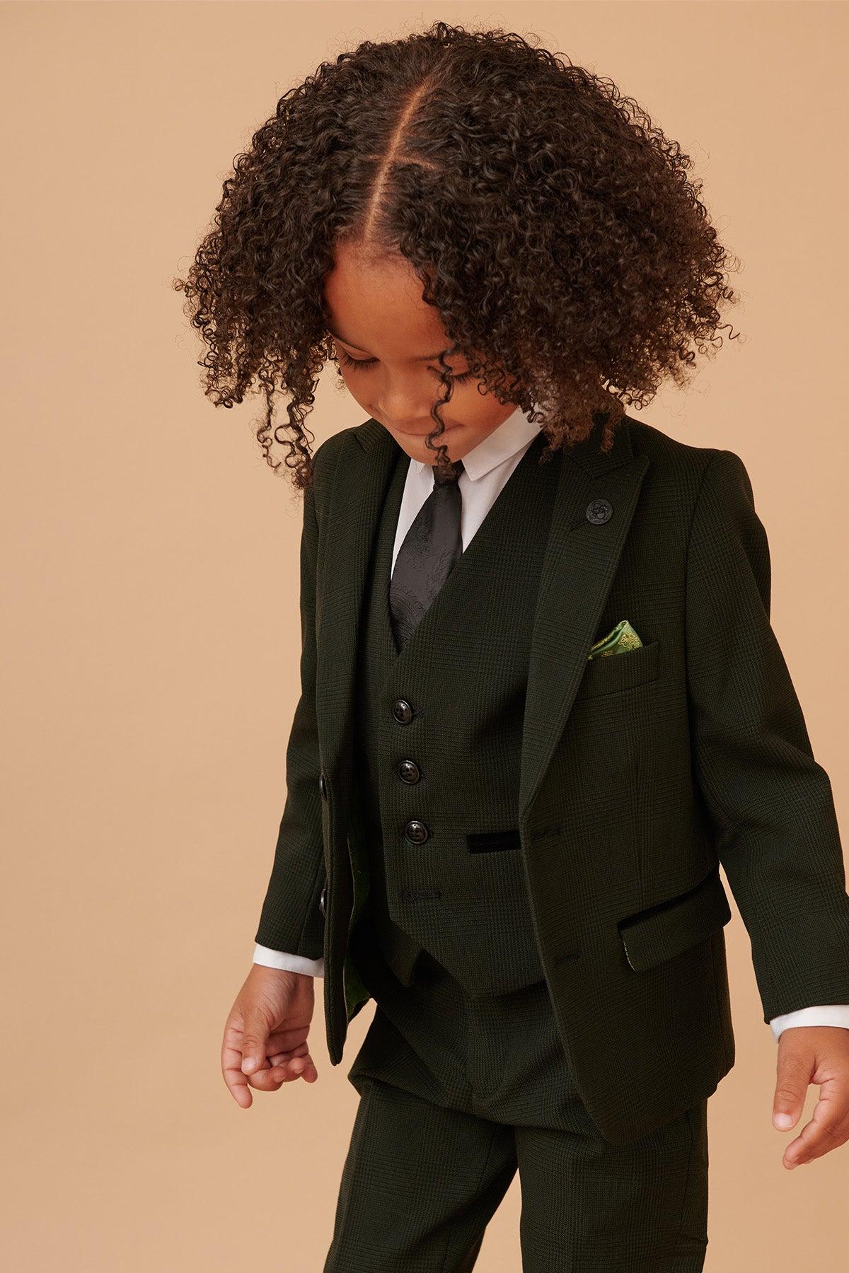 A young child models the BROMLEY - Childrens Olive Green Check Print Three Piece Suit by Marc Darcy, featuring a matching vest, white shirt, black tie, and green pocket square against a beige background.