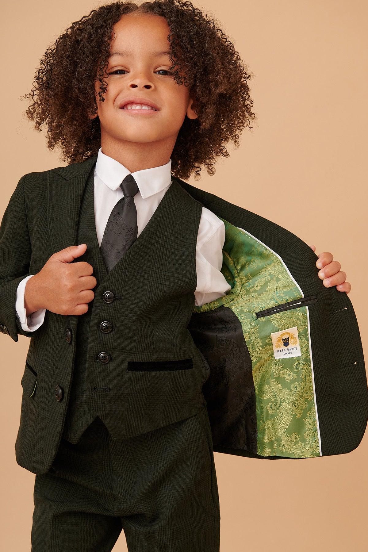 A smiling young child in the Marc Darcy BROMLEY children’s olive green check print three piece suit opens the jacket to show its green patterned lining and yellow label, styled with a white shirt and black tie.