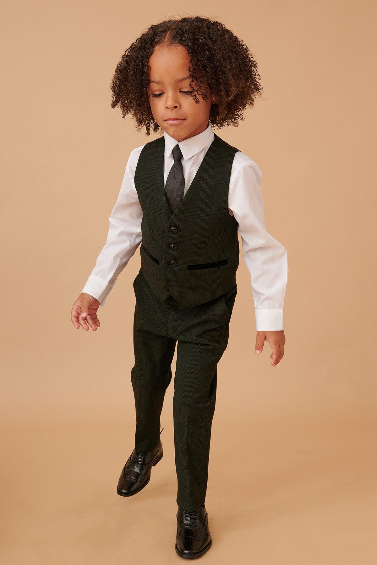 A young boy with curly hair wears the Marc Darcy BROMLEY childrens olive green check print three piece suit, featuring a vest and matching pants, paired with a white shirt and black tie, standing against a beige background.