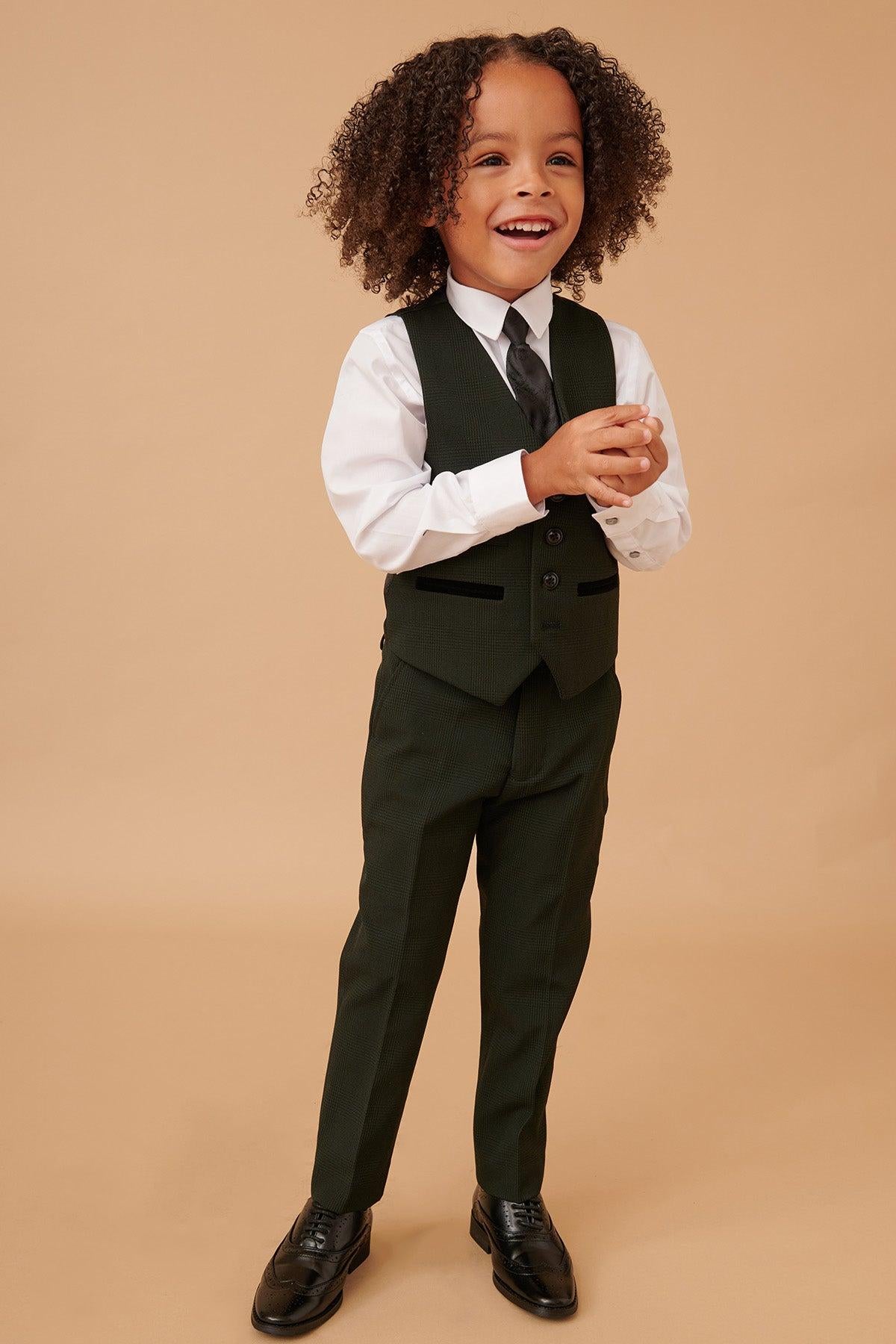 A young Black boy with curly hair smiles widely while wearing the Marc Darcy BROMLEY Children's Olive Green Check Print Three Piece Suit, paired with a white shirt and black tie.