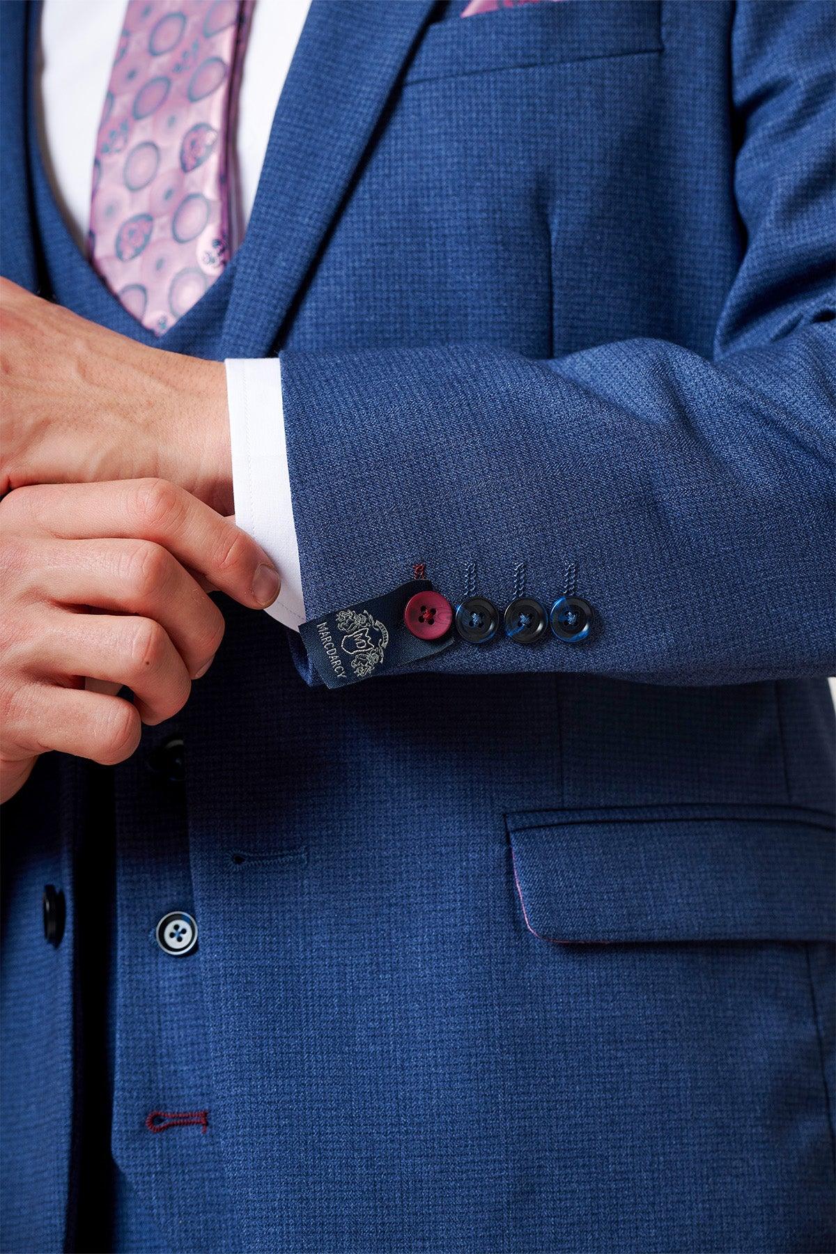 Close-up of the ASHLEY - Navy Blue Two Piece Suit by Marc Darcy: a textured slim fit jacket. His hand adjusts the crisp white shirt cuff. The sleeve has four buttons—one red, three black—and a dark navy Marc Darcy label.