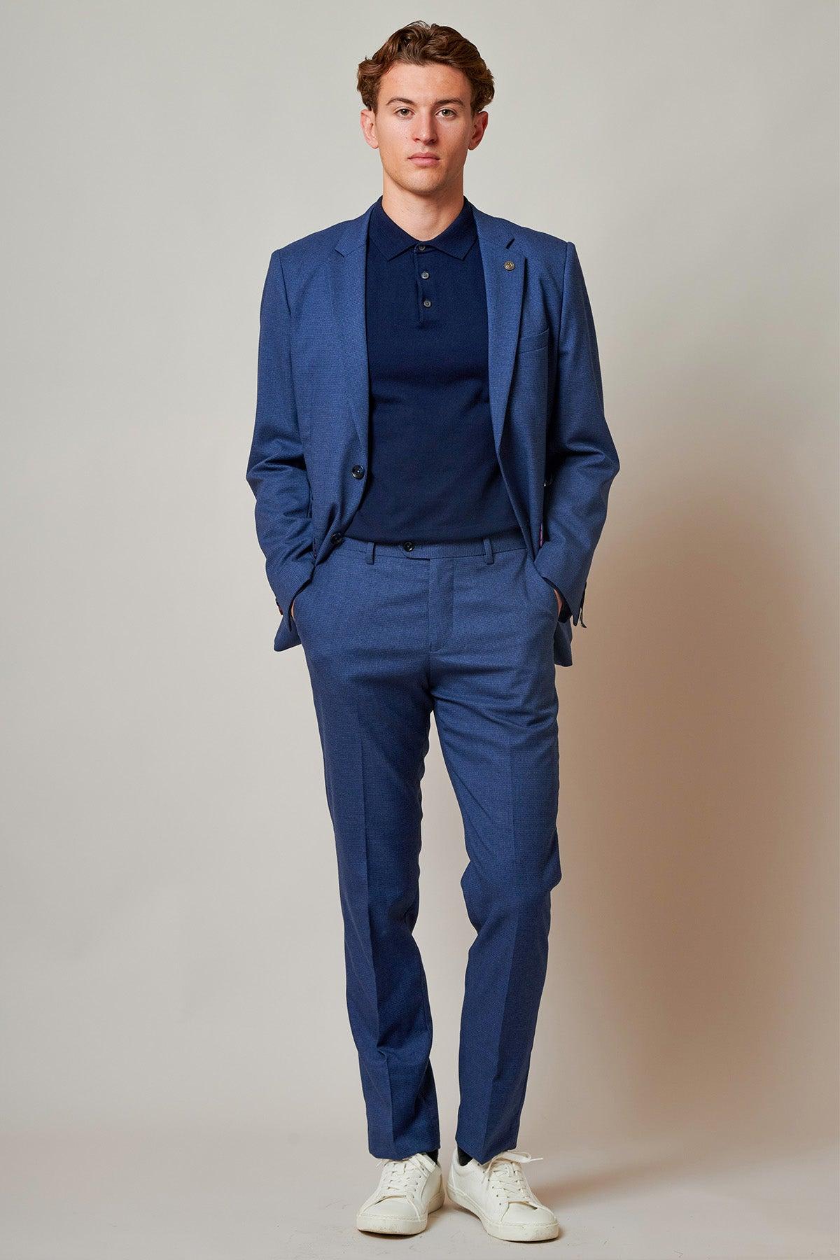 A young man stands against a plain backdrop wearing the Marc Darcy ASHLEY - Navy Blue Two Piece Suit with a navy polo shirt and white sneakers, hands in his pockets, looking straight ahead.