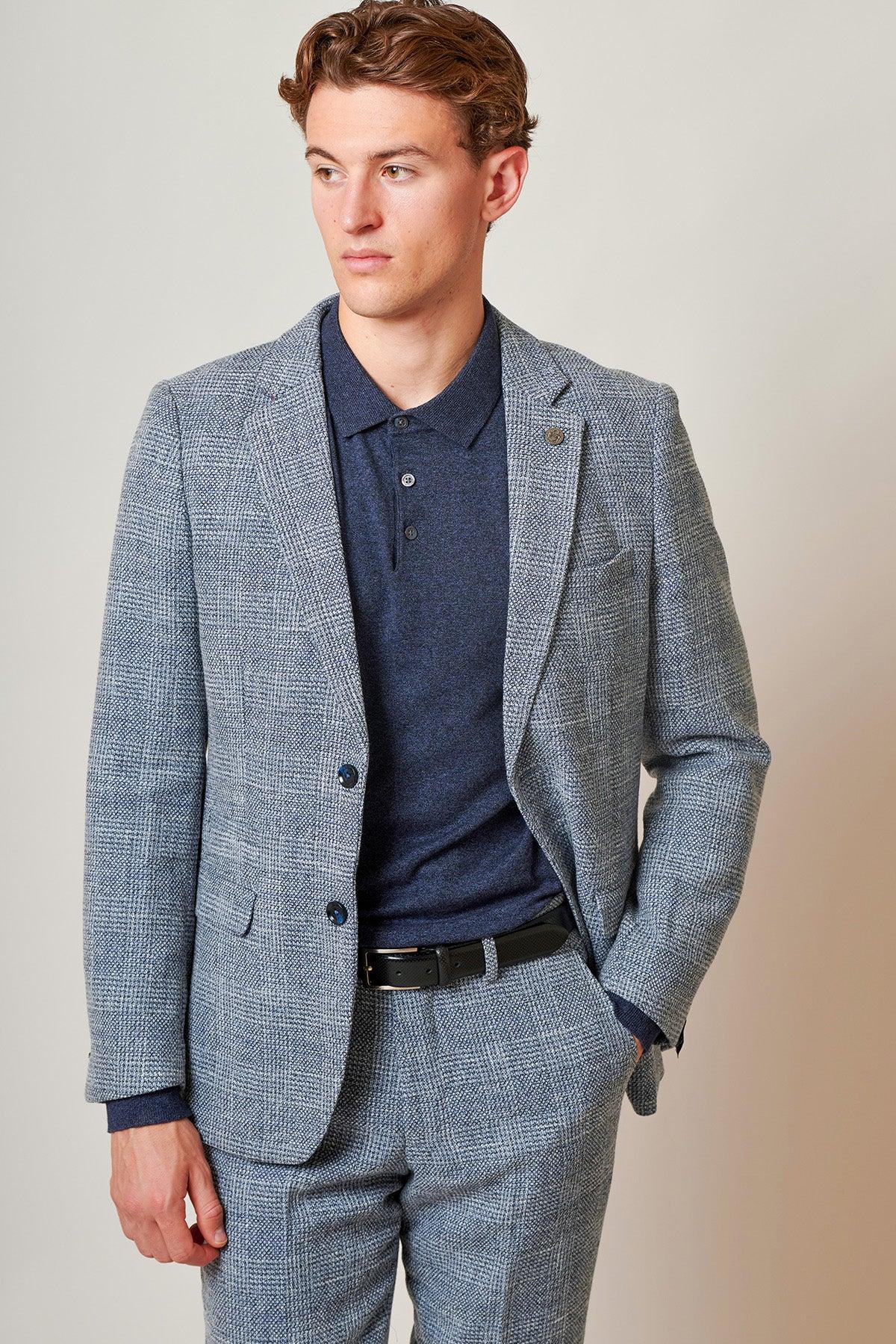 Tim Iroegbunam wears the KRIS Sky Blue Tweed Suit from The Everton Collection by marcdarcy, paired with a navy polo and black belt, standing with one hand in his pocket against a plain light background.