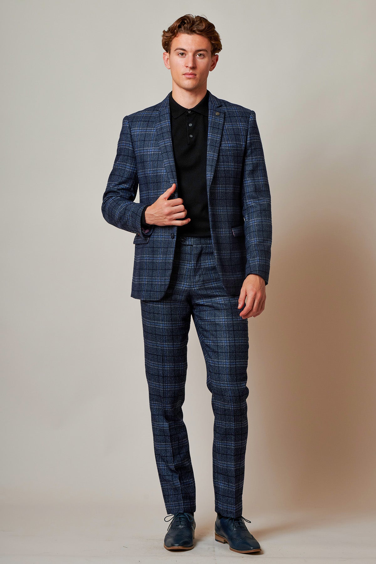 A man with short brown hair and a calm expression stands against a plain light background wearing the Marc Darcy ANDY - Blue Check Two Piece Suit, paired with matching trousers, a black polo shirt, and blue dress shoes, one hand raised to his chest.