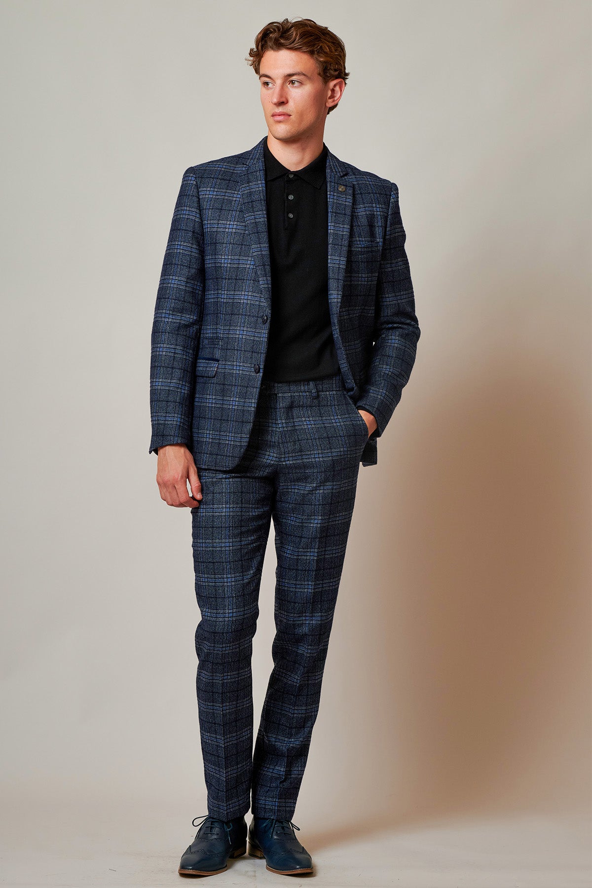 A man models the Marc Darcy ANDY Blue Check Two Piece Suit, styled with a black collared shirt and dark shoes. He stands against a plain background, left hand in pocket, gazing to the side with a neutral expression.