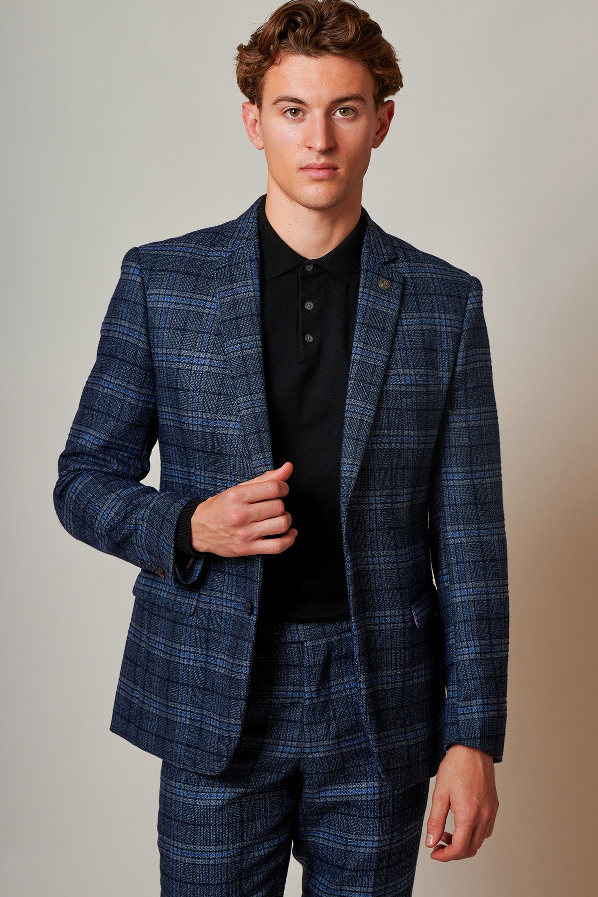 A young man wears the Marc Darcy ANDY Blue Check Two Piece Suit with a black polo shirt, lightly touching the lapel of his jacket.